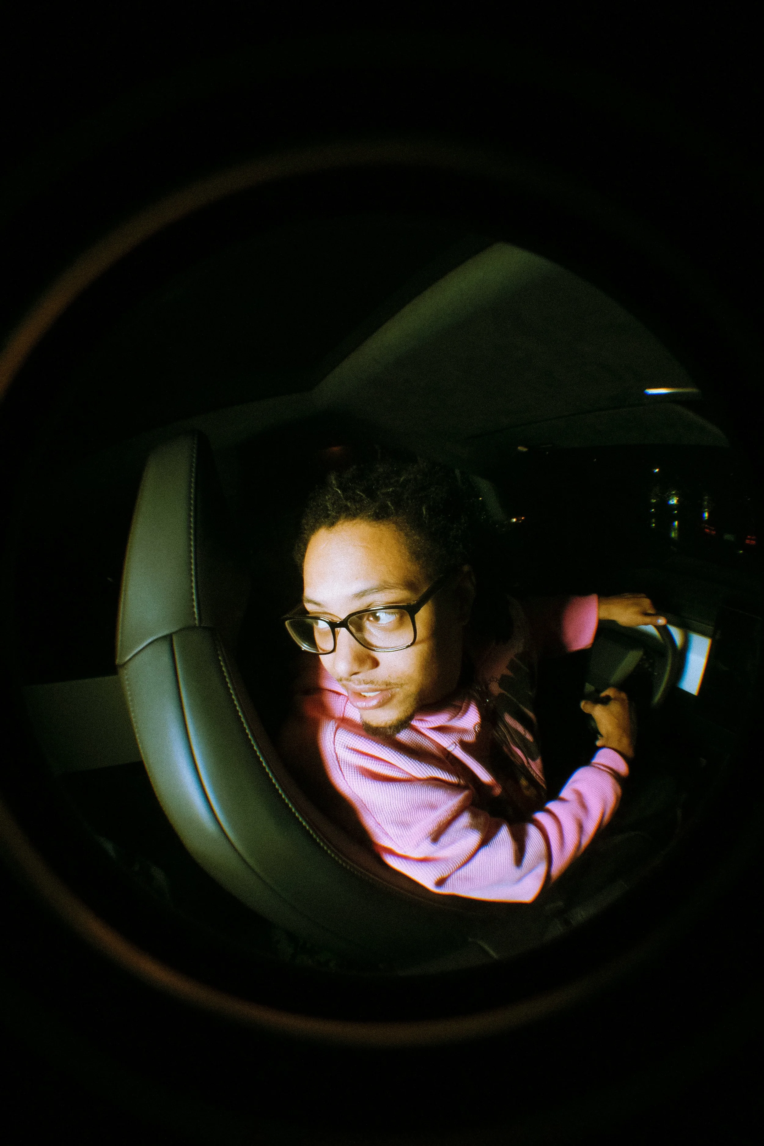 A person with glasses and curly hair sitting in the backseat of a car, looking out of the window, with a seatbelt on, during nighttime.