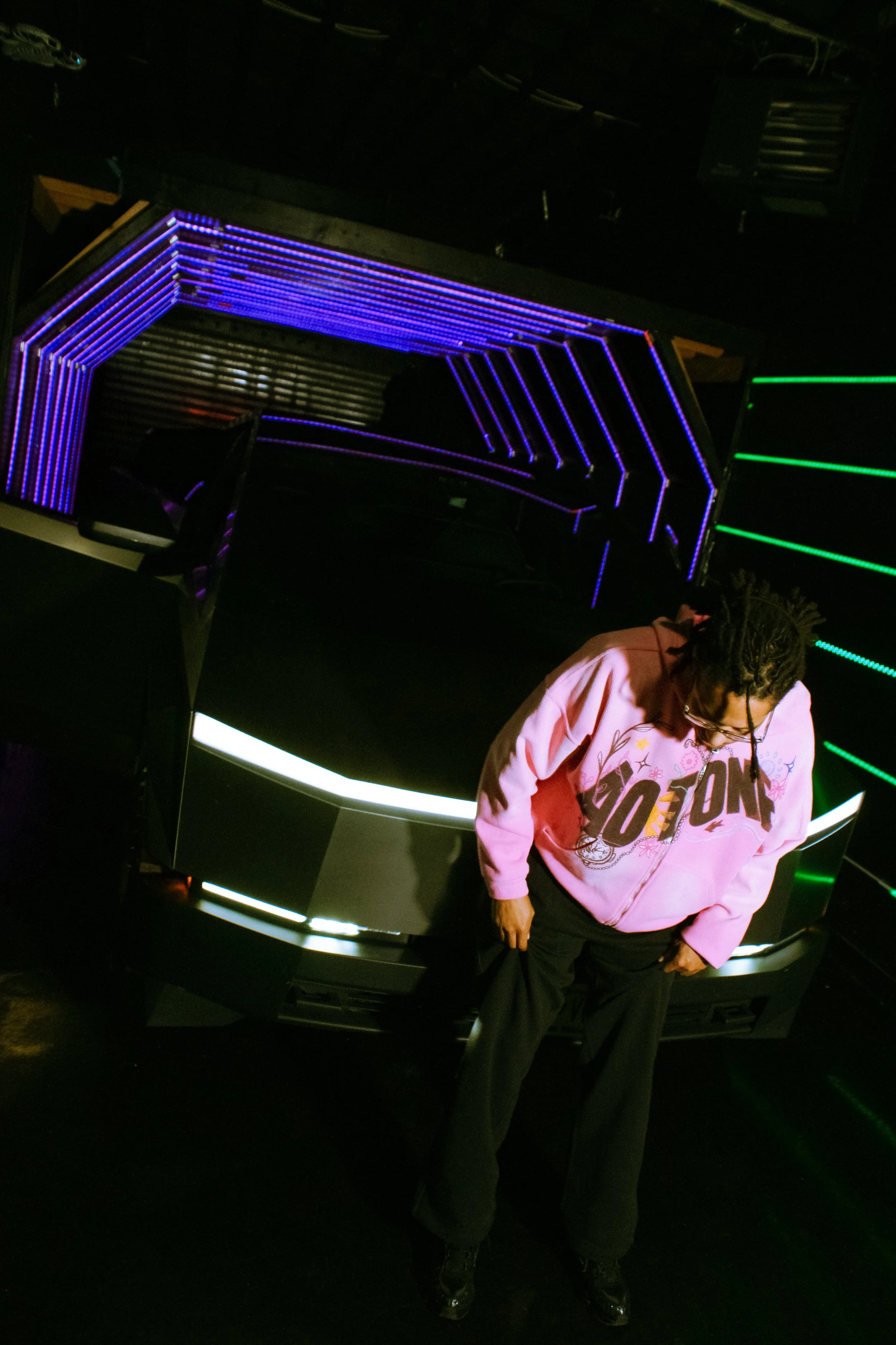 A woman with curly hair wearing a pink jacket and black pants, standing in front of a car with neon purple and green lights.