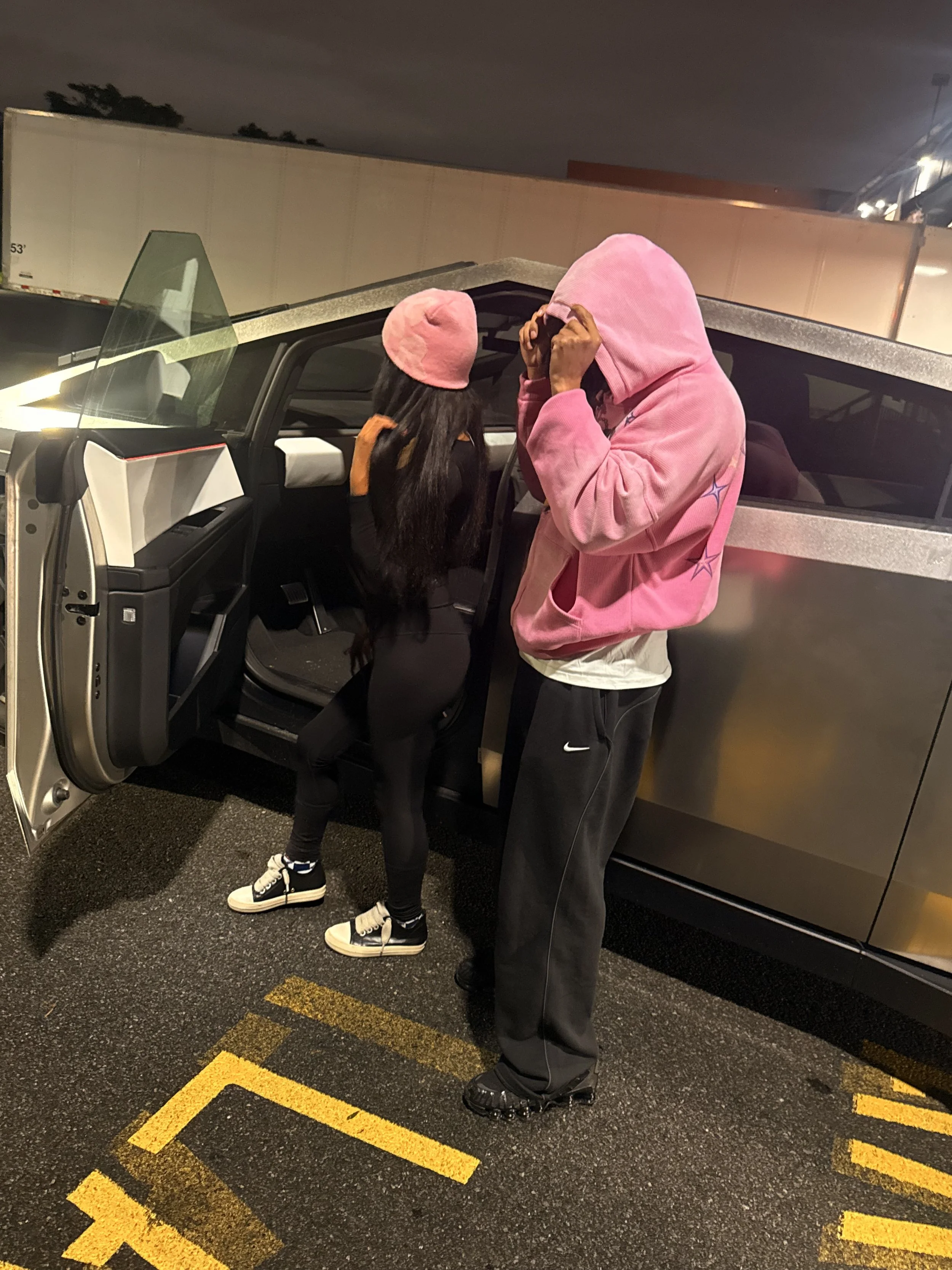 A person in a pink hoodie, standing next to a woman in a pink hat and black outfit, near an open car door at night in a parking lot.