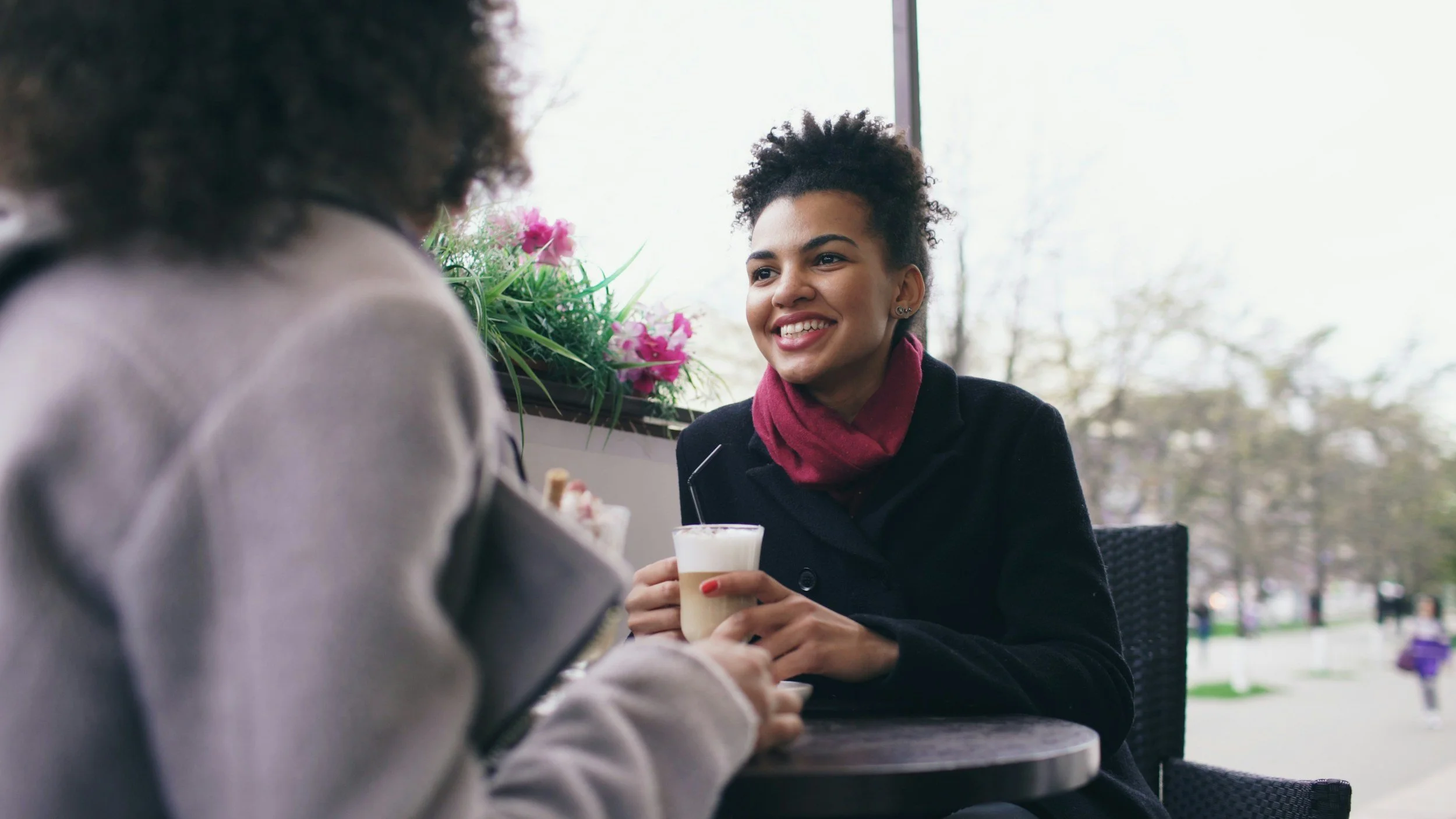 A woman with short curly hair and earrings smiling while holding a glass of a creamy beverage. She is sitting at an outdoor cafe table, conversing with another person whose blurred figure is visible in the foreground, near a potted plant with pink flowers, with trees and a park visible outside.