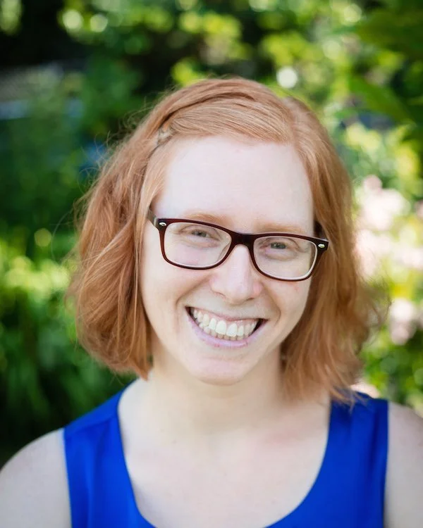 A woman with red hair, wearing glasses and a blue top, smiling outdoors with green foliage in the background.