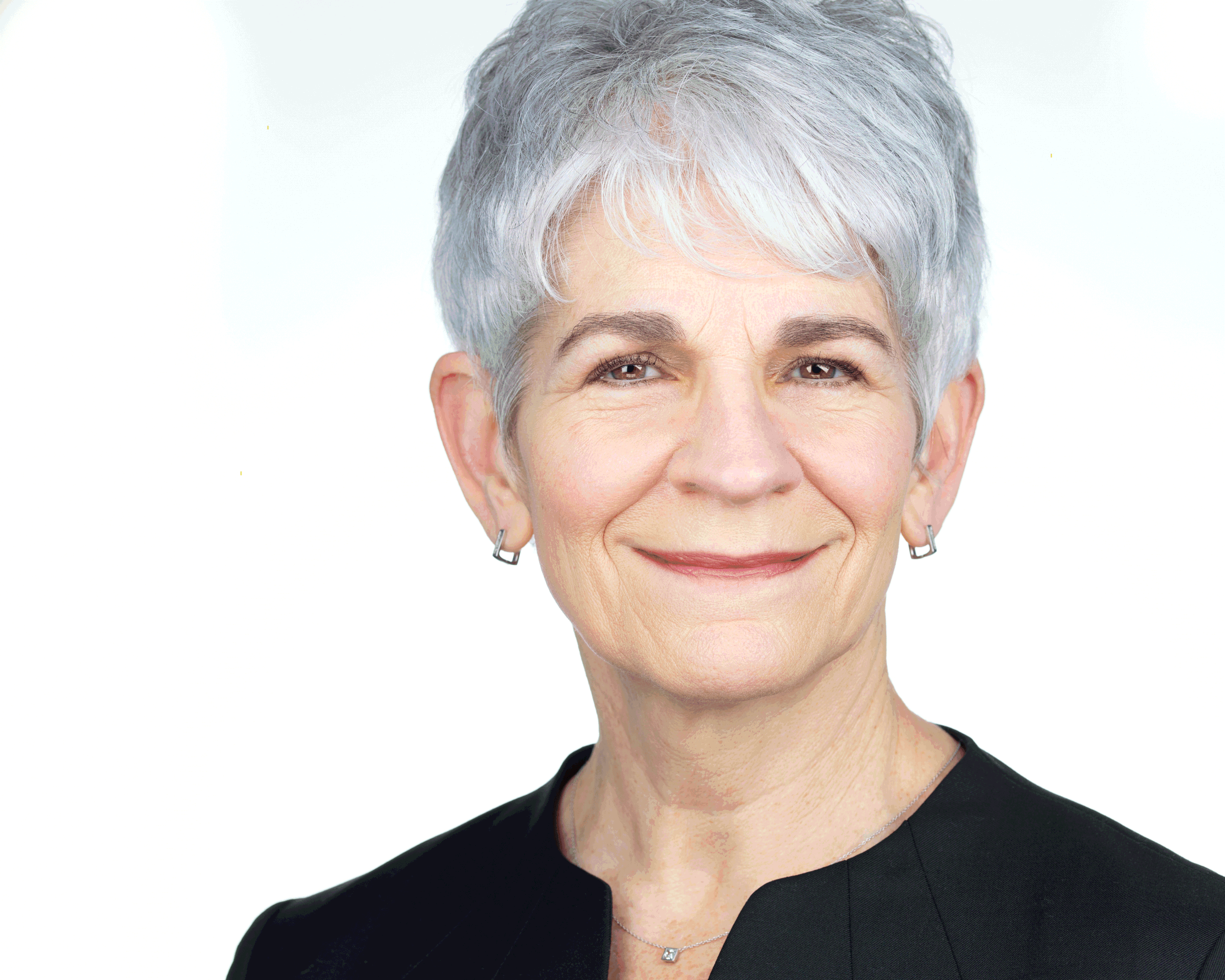 Close-up of a smiling older woman with short gray hair, wearing earrings and a black top, against a white background.
