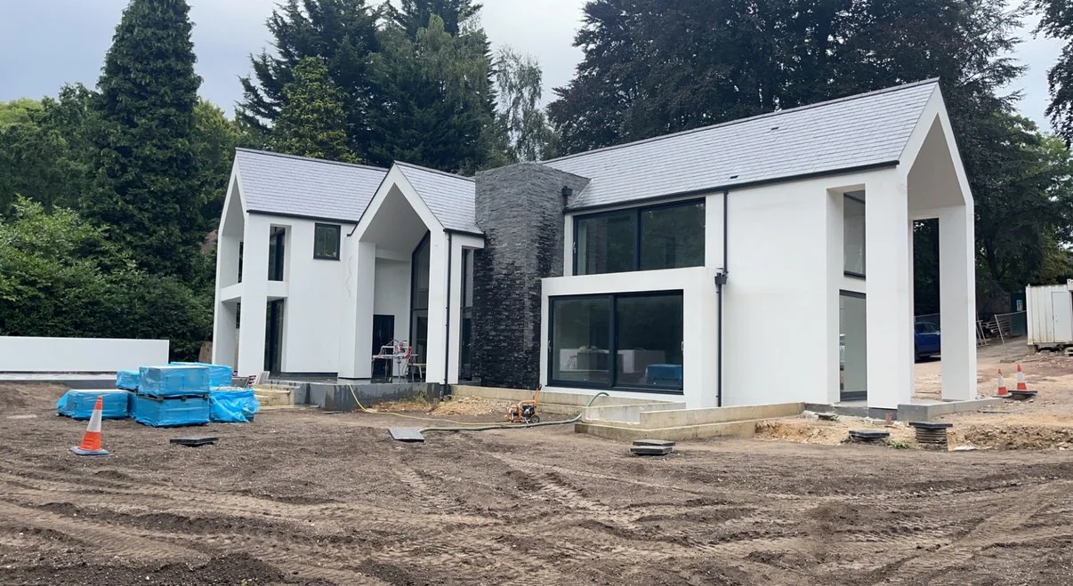 Under construction modern two-story white house with black window frames, large glass windows, and a central gray stone chimney, set against a backdrop of green trees.