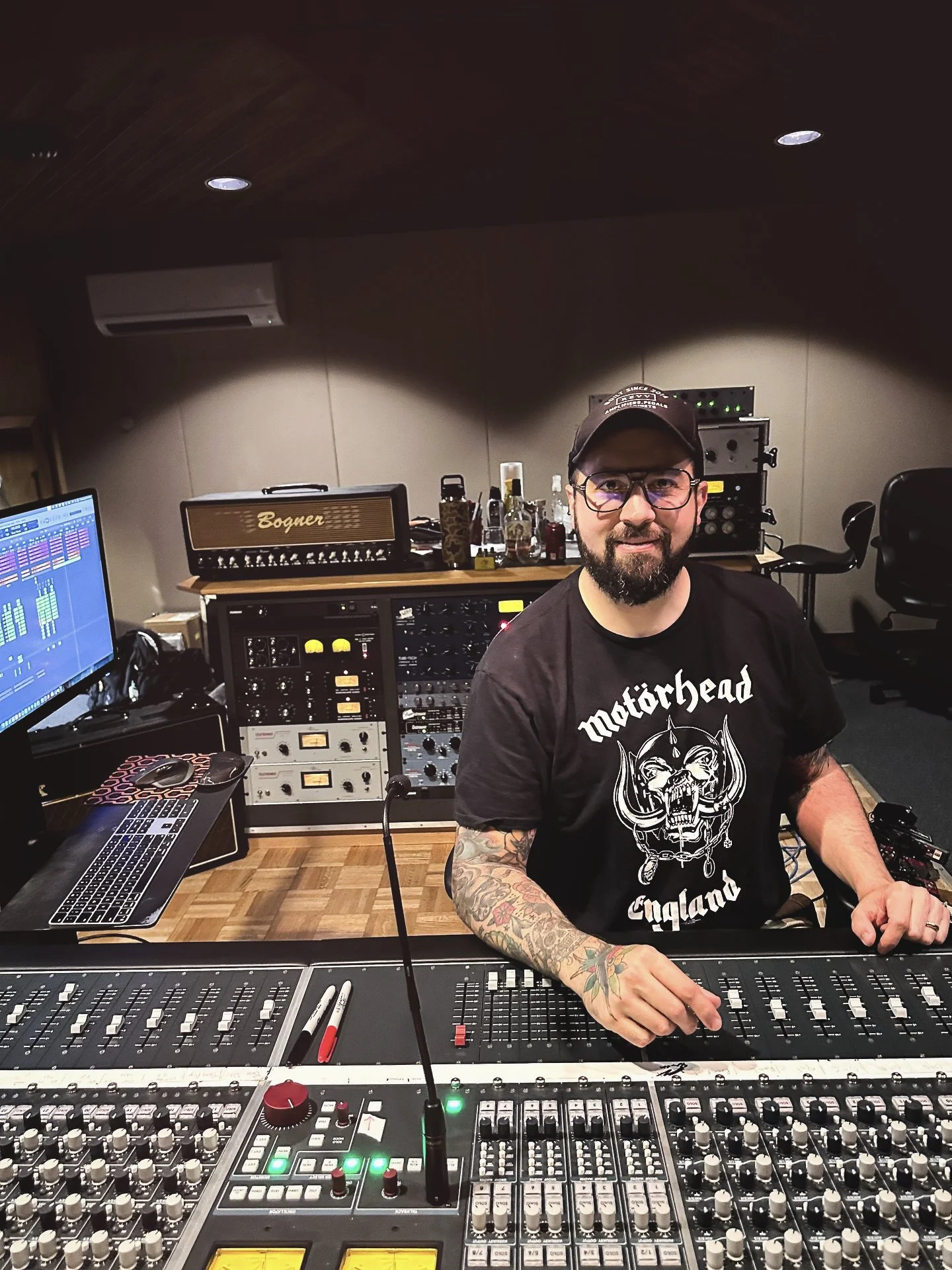 A man with glasses and tattoos on his arm sitting at a mixing console in a recording studio, wearing a Motorhead T-shirt and a cap. There are audio equipment, bottles, and a computer monitor in the background.