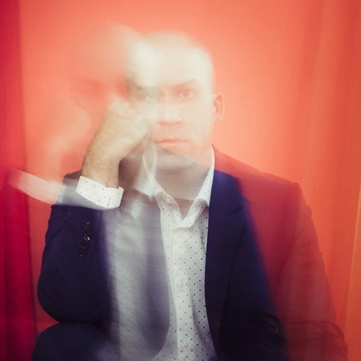 A man with short hair and a serious expression, wearing a white shirt with a small dotted pattern and a dark blazer, is seen through a glass or plastic surface with red and orange coloring, creating a blurred, layered effect.