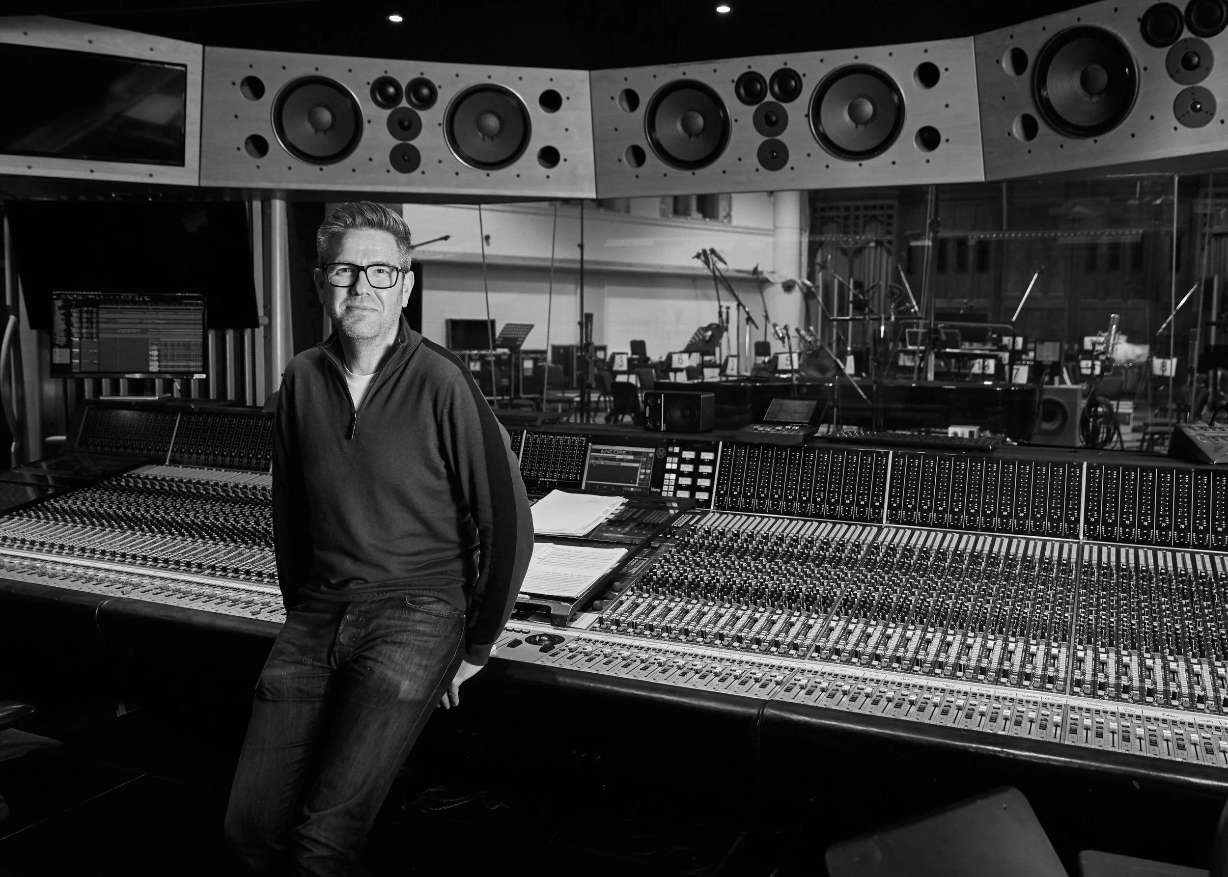 A man in glasses and a dark jacket standing in a music recording studio with large mixing console and multiple speakers in the background.