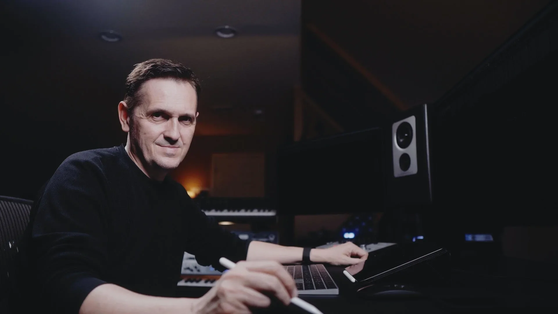 Man sitting at a digital music workstation in a dark studio, with a keyboard and large speaker.