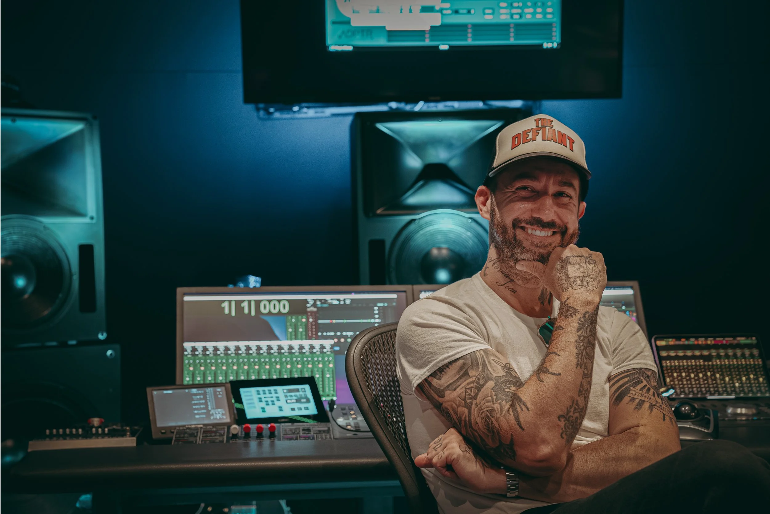 A man with tattoos wearing a beige cap with the words 'The Defiant', smiling in a music studio surrounded by sound equipment and computer screens.