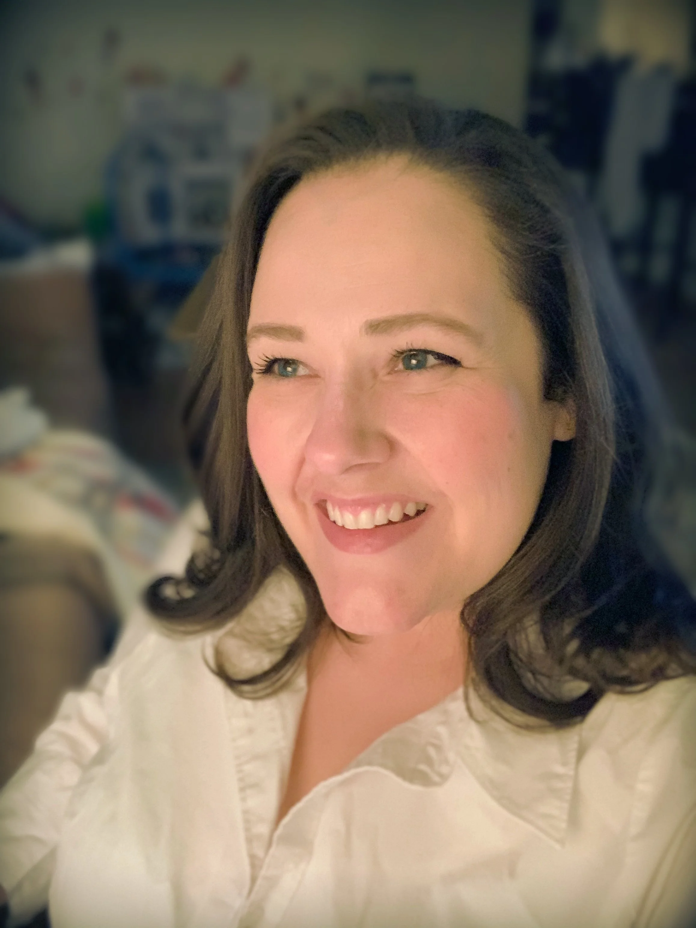Jen Aalgaard-Bretz, Founder & Principal Partner  | A smiling woman with shoulder-length brown hair and blue eyes, wearing a white shirt, in a blurred indoor setting.