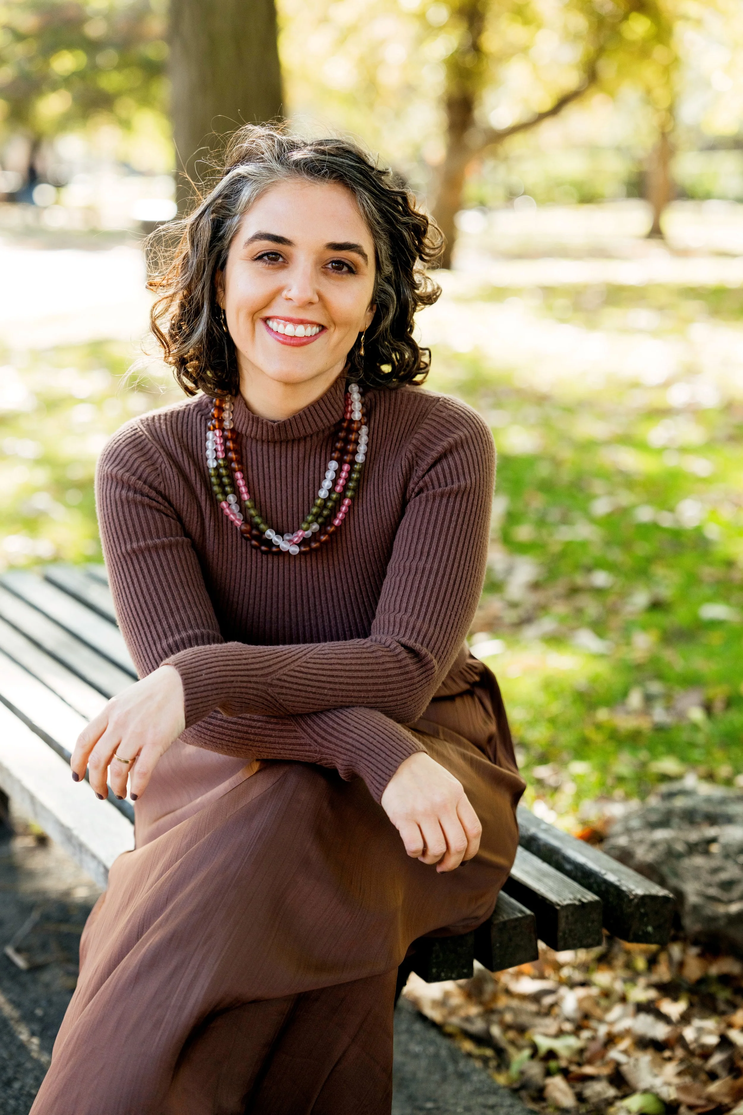 A woman with curly dark hair and light skin, sitting on a park bench outdoors, smiling at the camera, wearing a brown long sleeve sweater, brown skirt, and layered multicolored beaded necklaces.