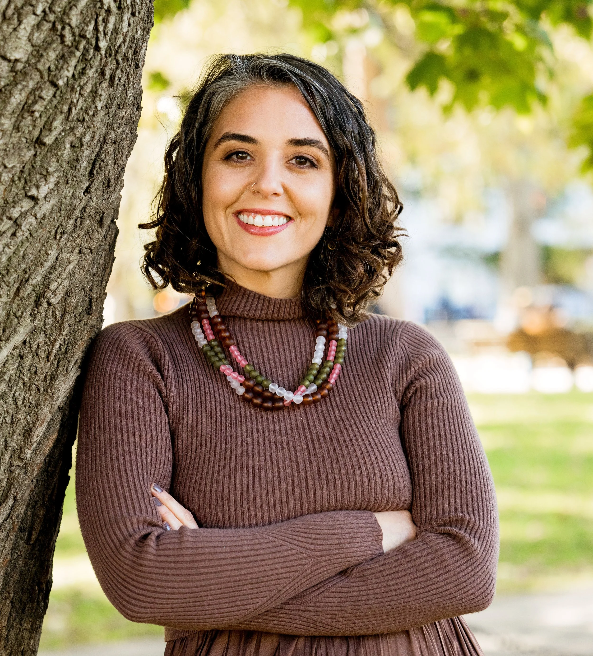 A woman with curly dark hair wearing a brown ribbed turtleneck dress and colorful beaded necklaces posing outdoors next to a tree with green leaves.