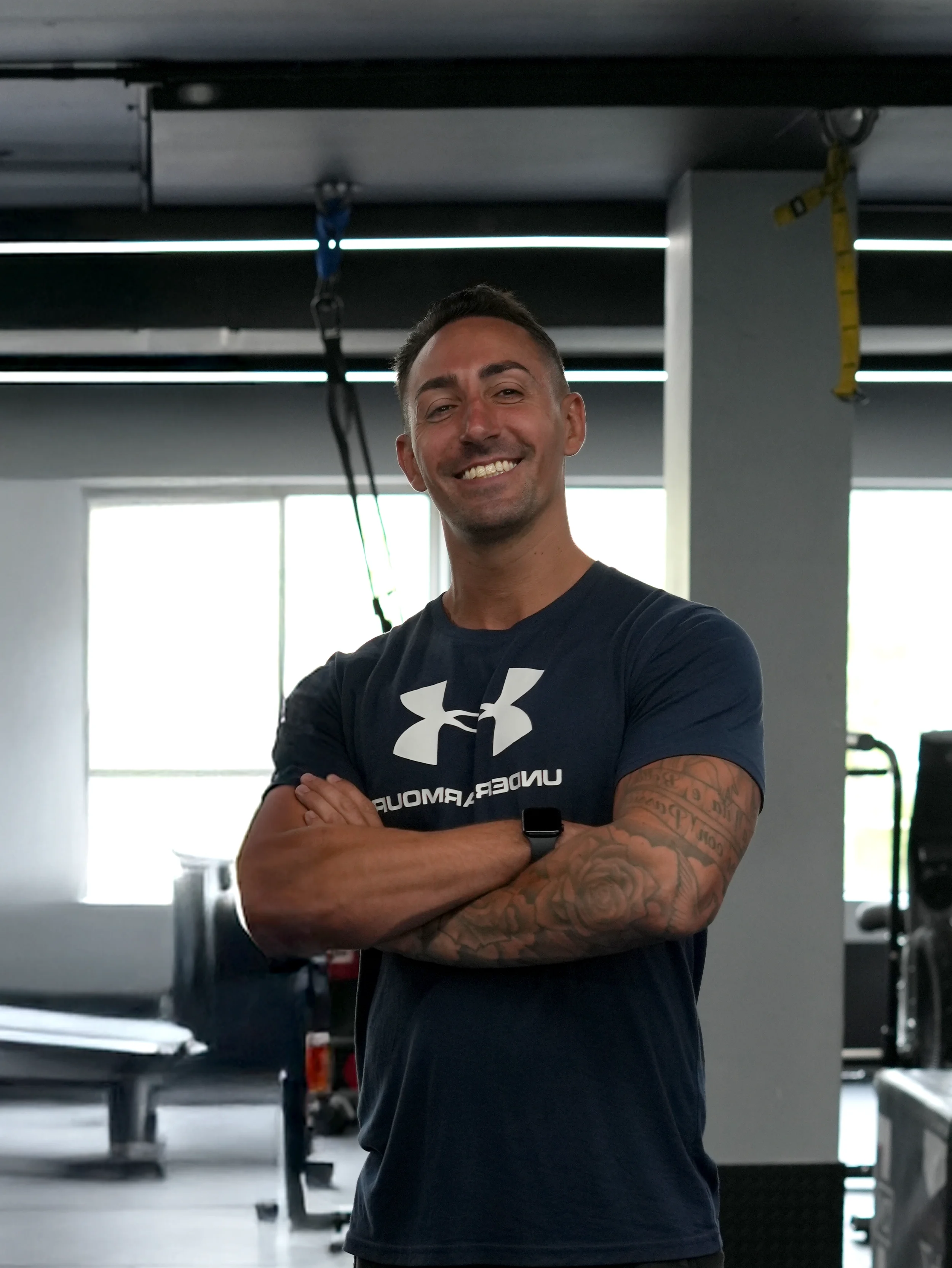 A smiling man with tattooed arms standing with arms crossed inside a gym, wearing a navy blue Under Armour t-shirt.