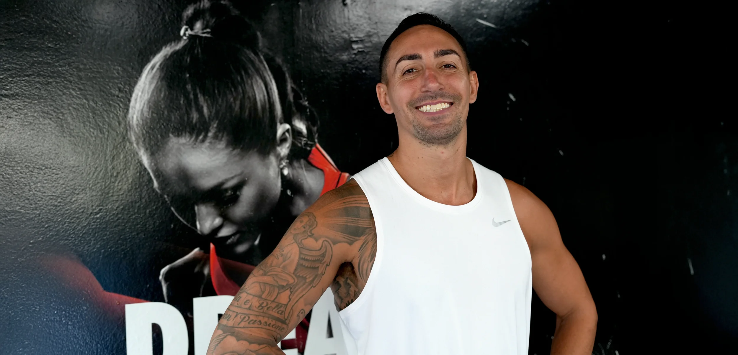 A smiling man with short dark hair and tattoos on his arm wearing a white athletic tank top standing in front of a black and white poster of a woman with braided hair, wearing a red top.