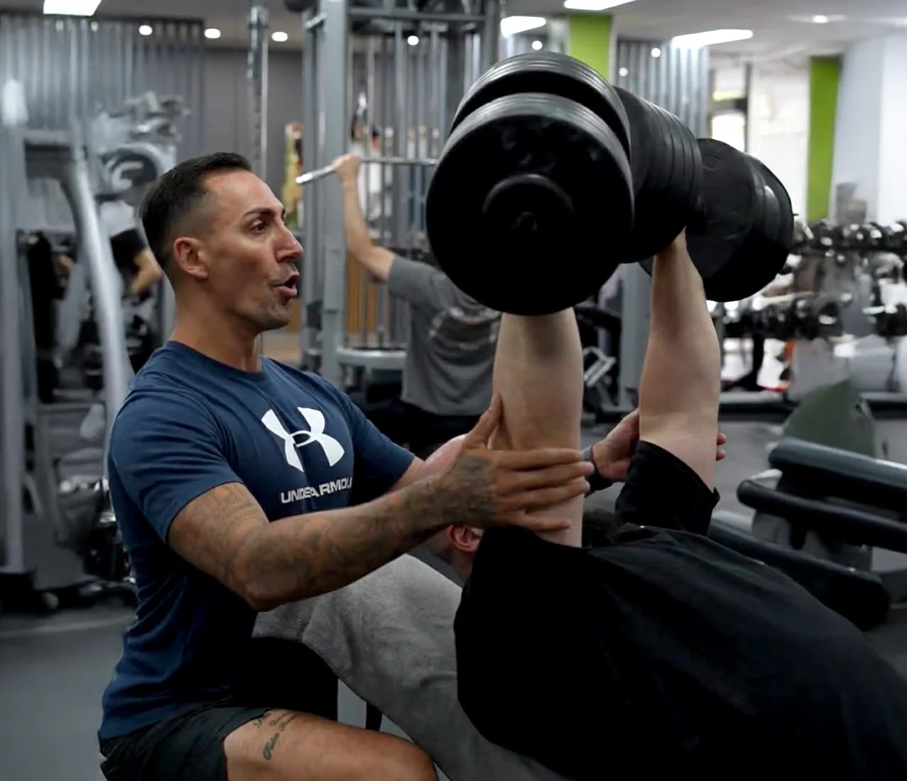 A male personal trainer assisting a client with a shoulder press exercise in a gym, with weights and workout equipment in the background.