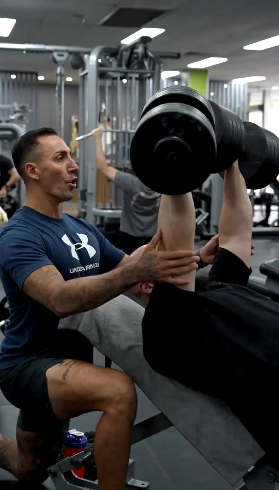 A personal trainer assisting a man with a seated shoulder press exercise at the gym.