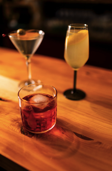 Three cocktails on a wooden bar counter: a martini with an olive, a champagne flute with lemon, and a negroni.