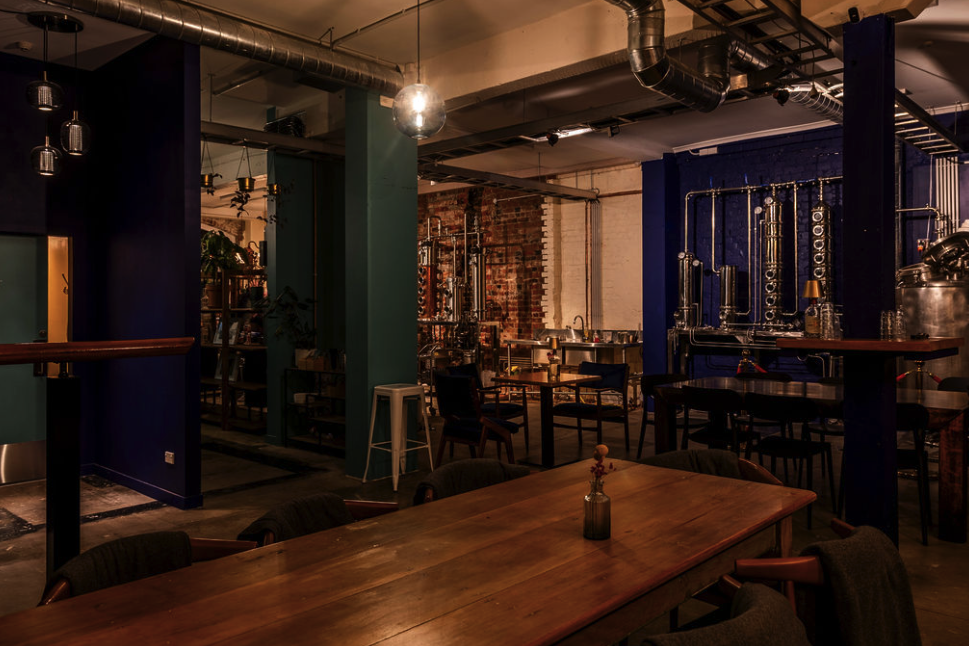 Interior of a modern, industrial-style bar with wooden tables, assorted chairs, exposed brick walls, and visible pipes and metallic fixtures.
