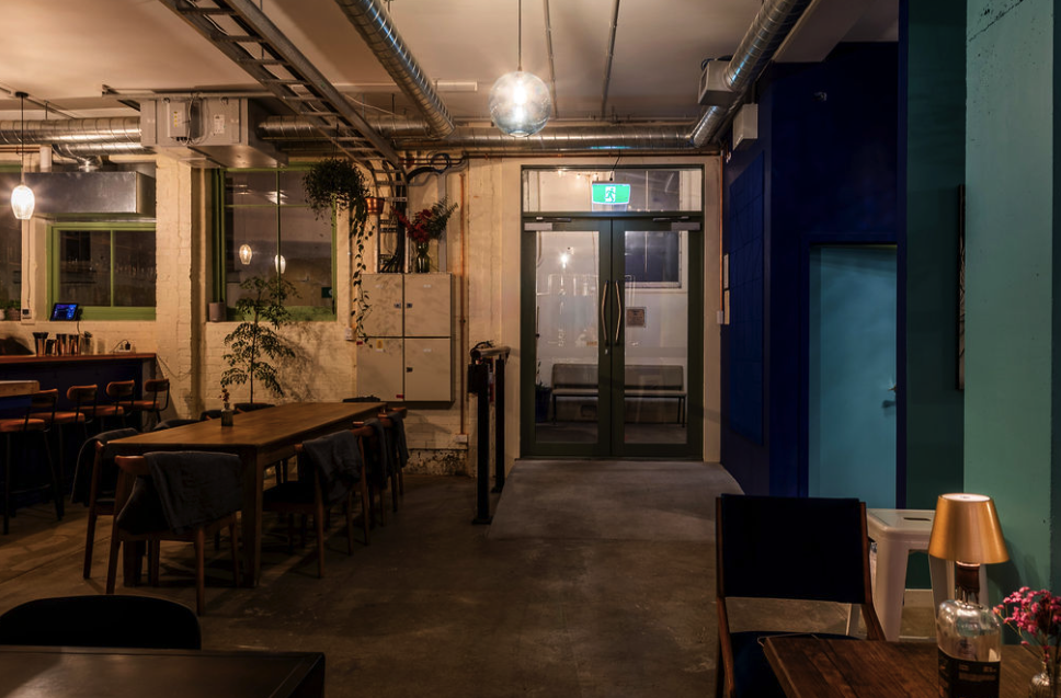 Interior of a modern industrial-style bar with wooden tables, chairs, indoor plants, and visible ductwork on the ceiling.