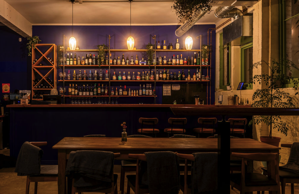 Interior of a cozy bar with a dark blue wall, wooden shelves filled with bottles of alcohol, pendant lights, potted plants, and a wooden dining table with chairs.