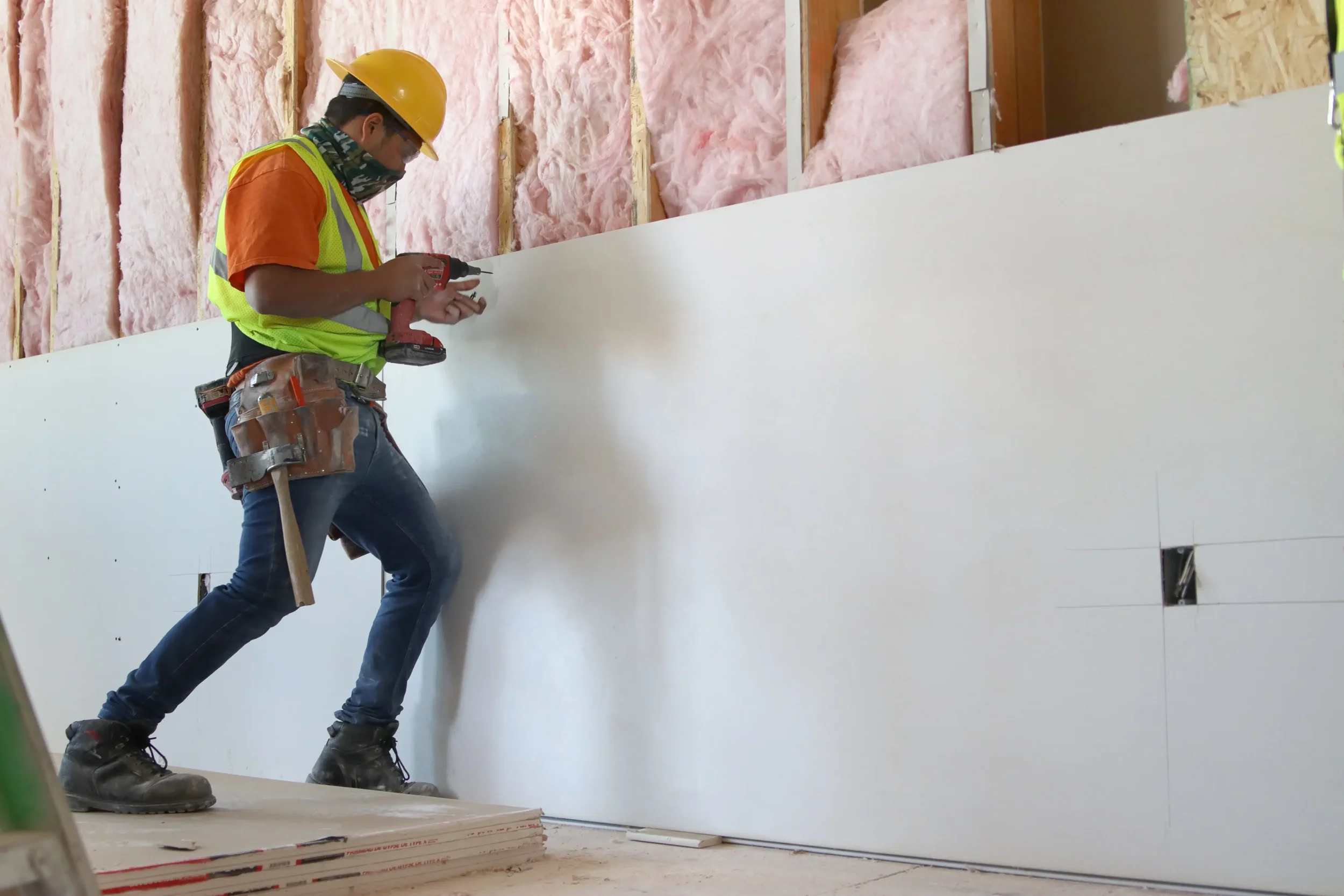 Construction worker wearing a yellow hard hat, safety vest, and face mask using a power drill to work on a drywall inside a building.