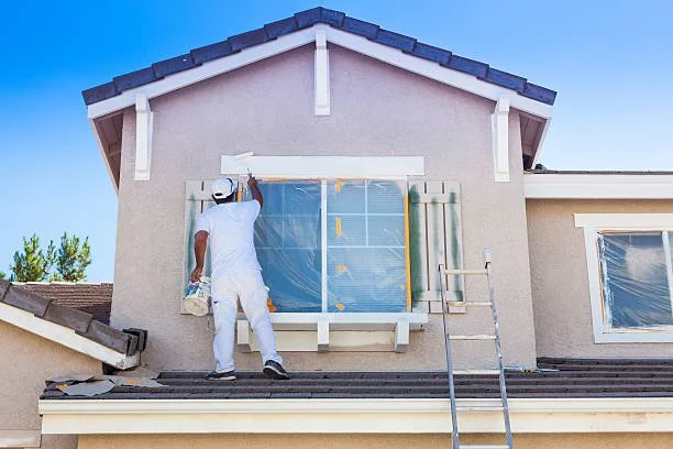 A painter in white clothes and a white cap painting a window frame on the exterior of a house. The house has beige walls and a dark tiled roof, with a ladder leaning against it. The window is covered with plastic for protection.
