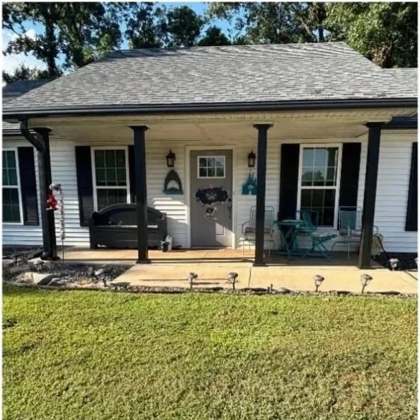Front porch of a house with black porch columns, white siding, and a small wooden deck. Outdoor furniture and decorations are visible, along with a lawn and trees in the background.