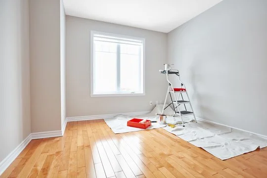 Empty room with hardwood floors, a window, and painting supplies including a step ladder, paint tray, and drop cloth.