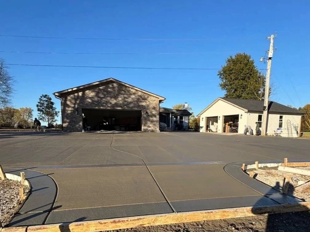 Newly poured concrete driveway with construction framing, two garage buildings, and a utility pole in a residential area.