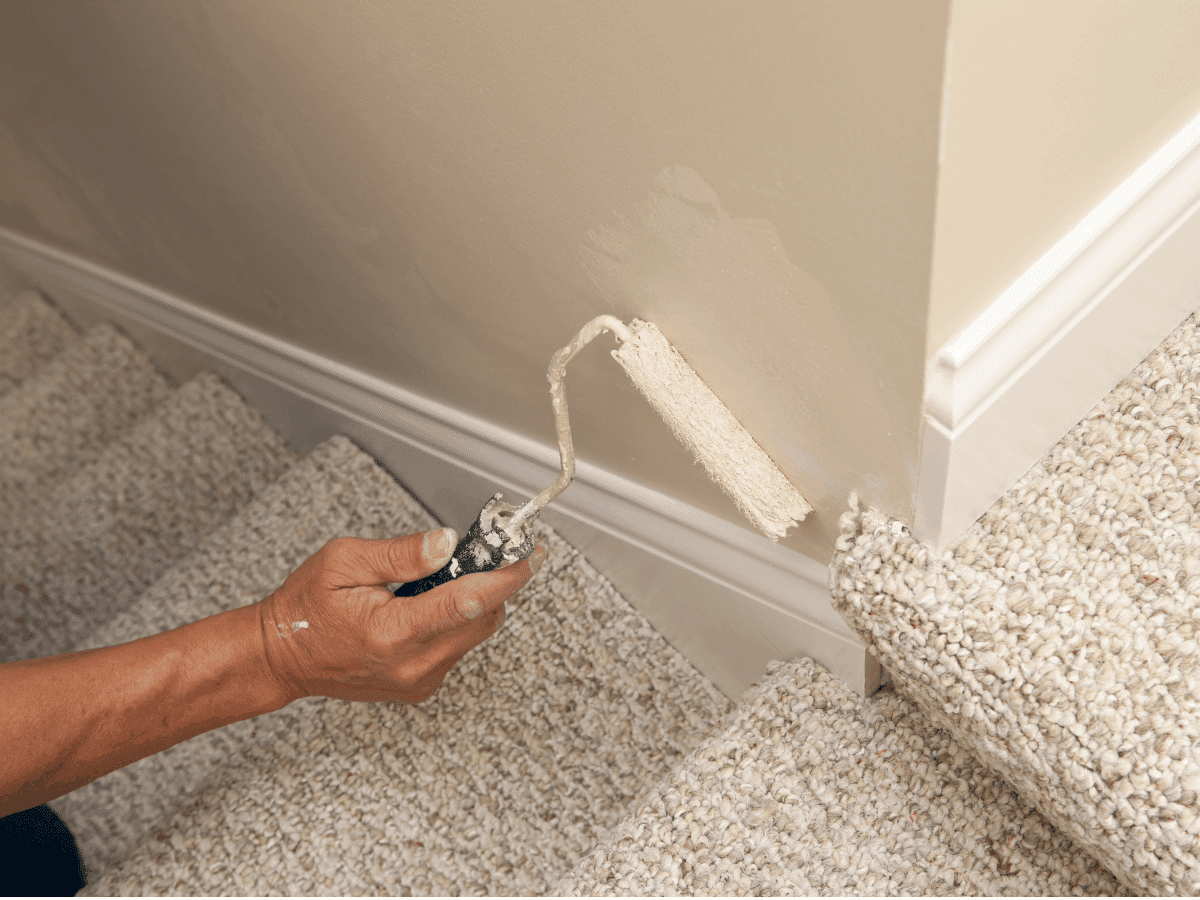 Person painting the baseboard with a roller brush near carpeted stairs.