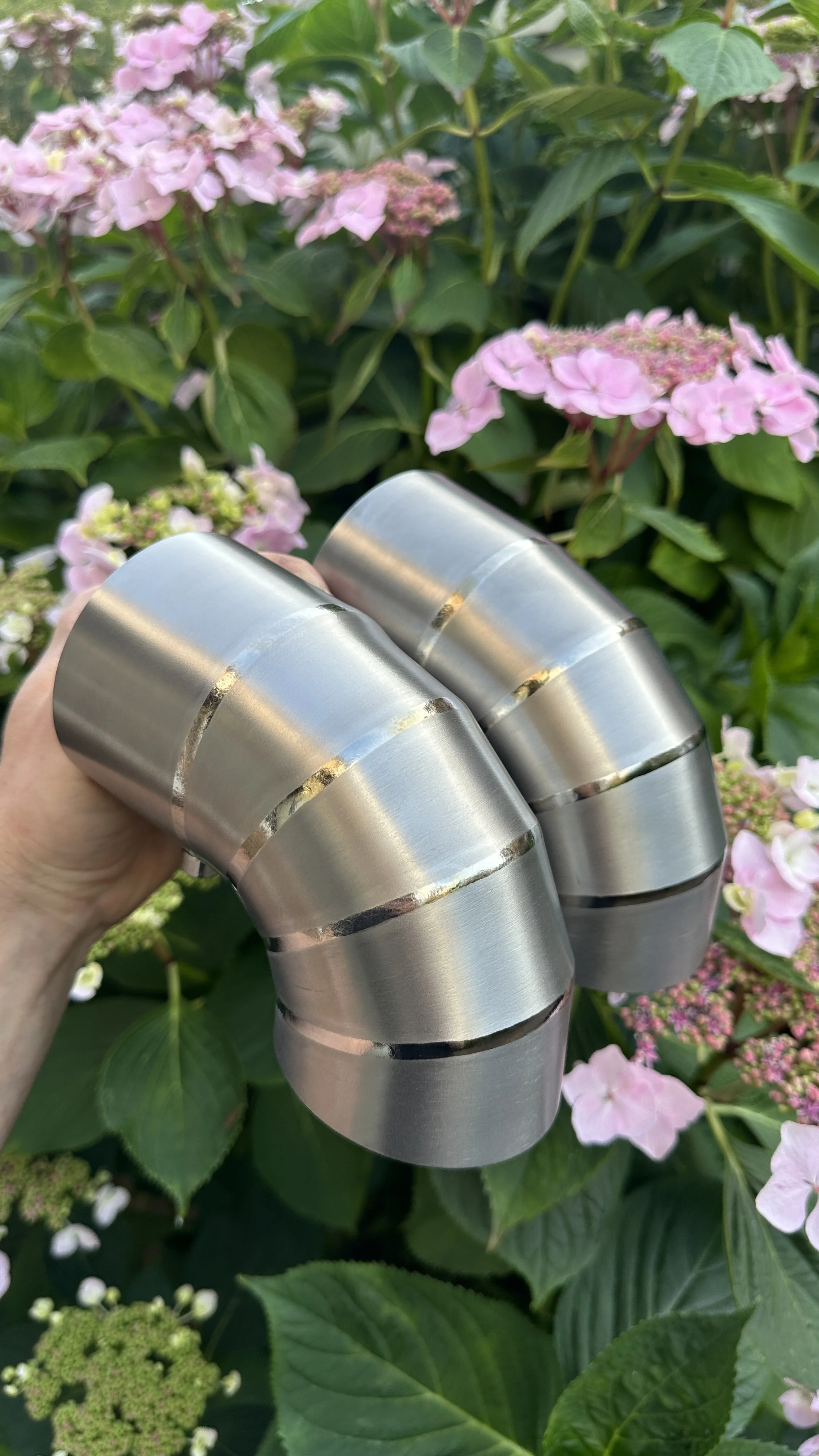 Close-up of two metallic, curved, segmented objects held in a hand against a background of pink hydrangea flowers and green leaves.