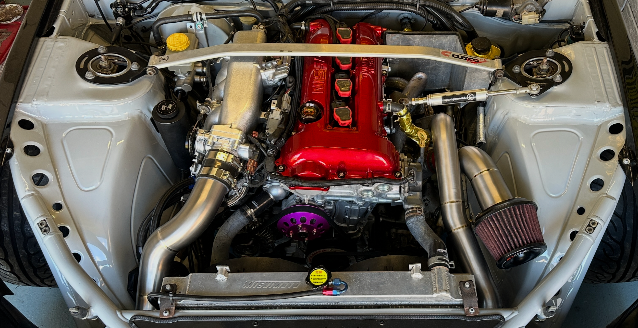 Close-up of a modified car engine with red valve cover, titanium intake pipes, air filter, and strut brace.