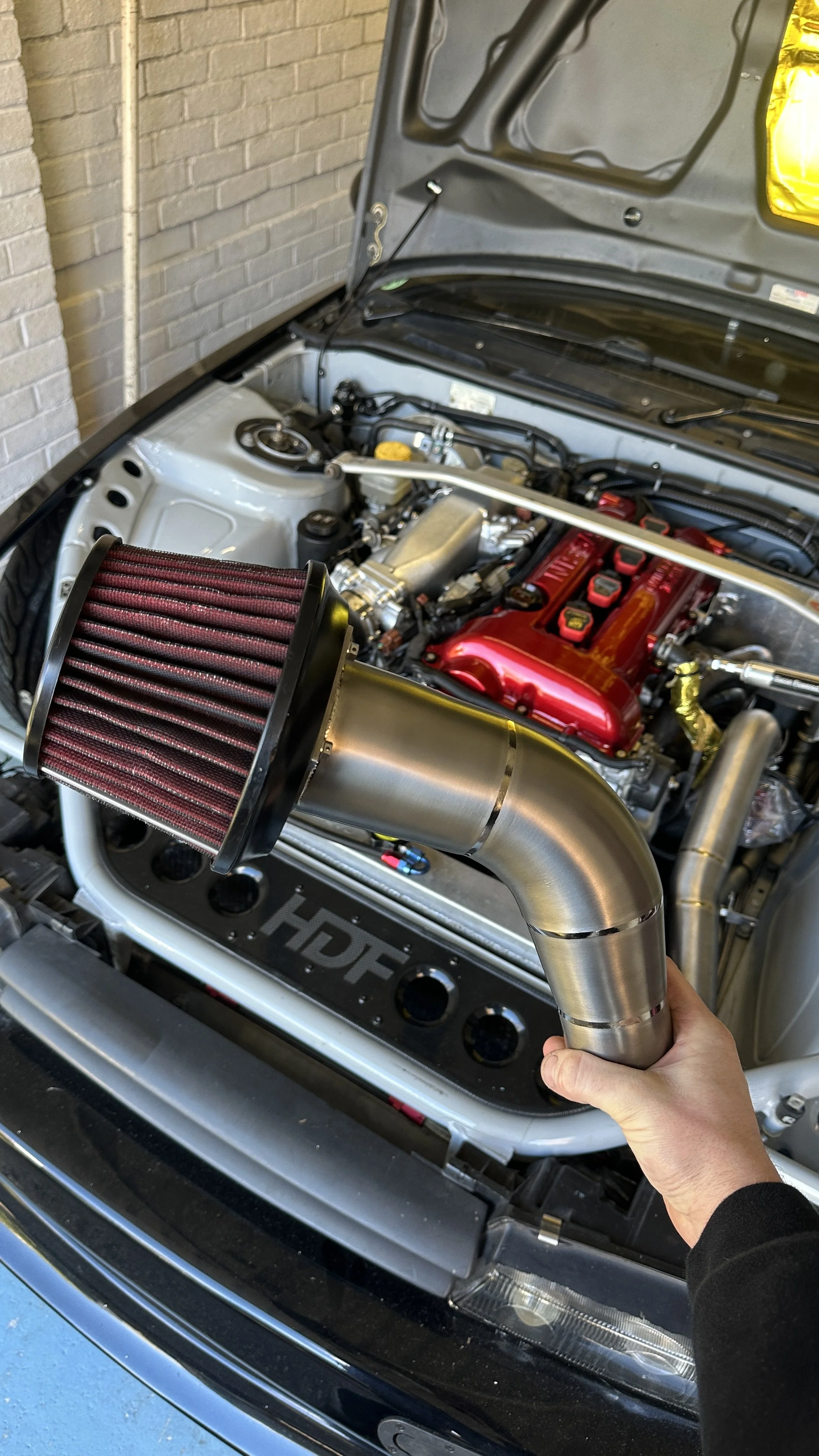 A person holding a large cold air intake pipe with a red and black air filter in front of a car engine bay.