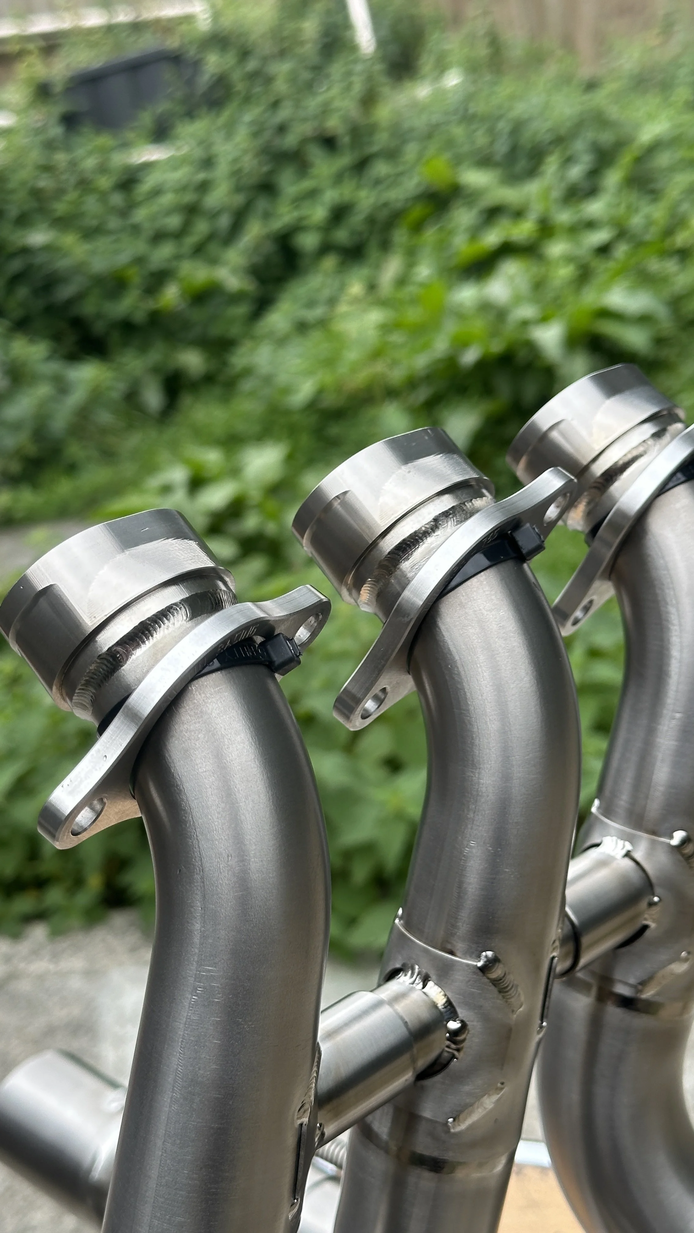 Close-up of three stainless steel bicycle handlebars with mounting brackets, set outdoors with greenery in the background.
