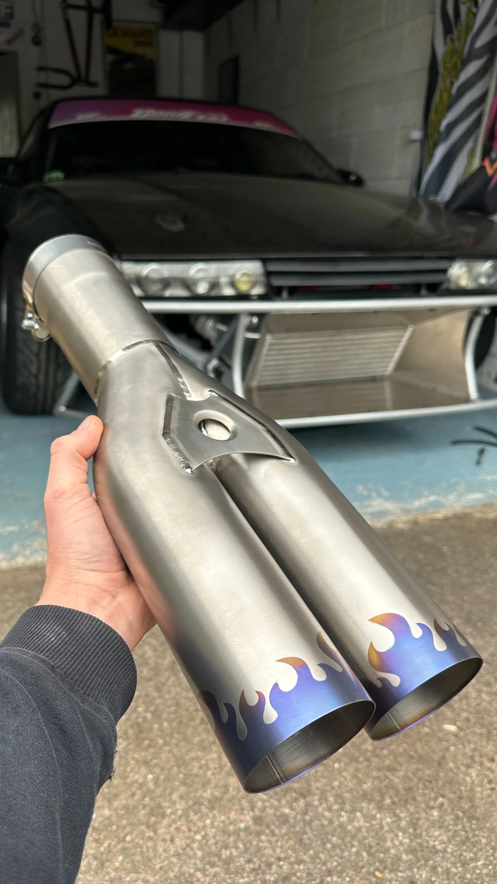 Person holding a metal exhaust pipe with flame design at the end, in front of a black race car with an open front grill.