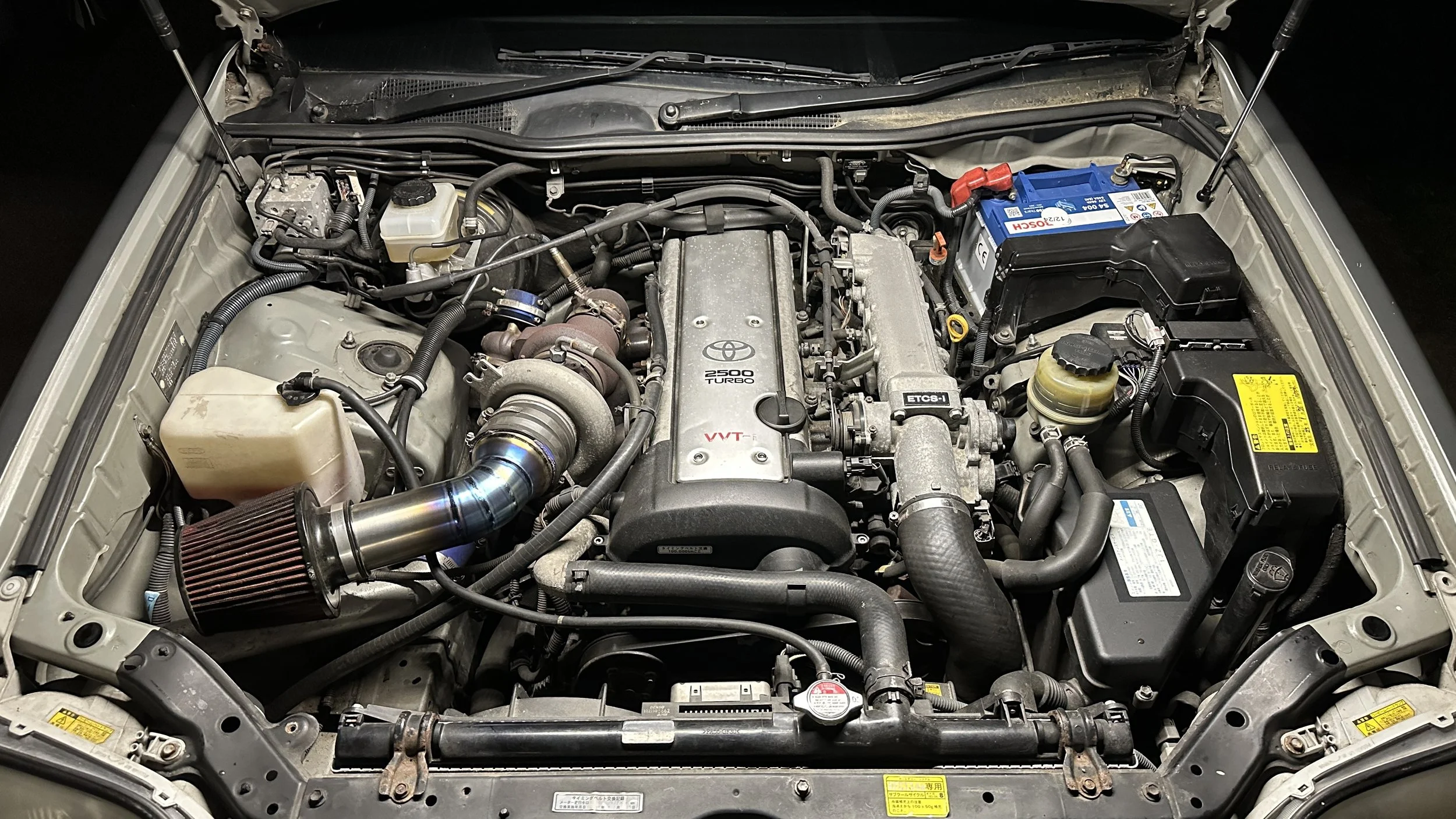 Under the hood of a car showing the engine compartment with a turbocharged engine, air intake, battery, and various engine components.