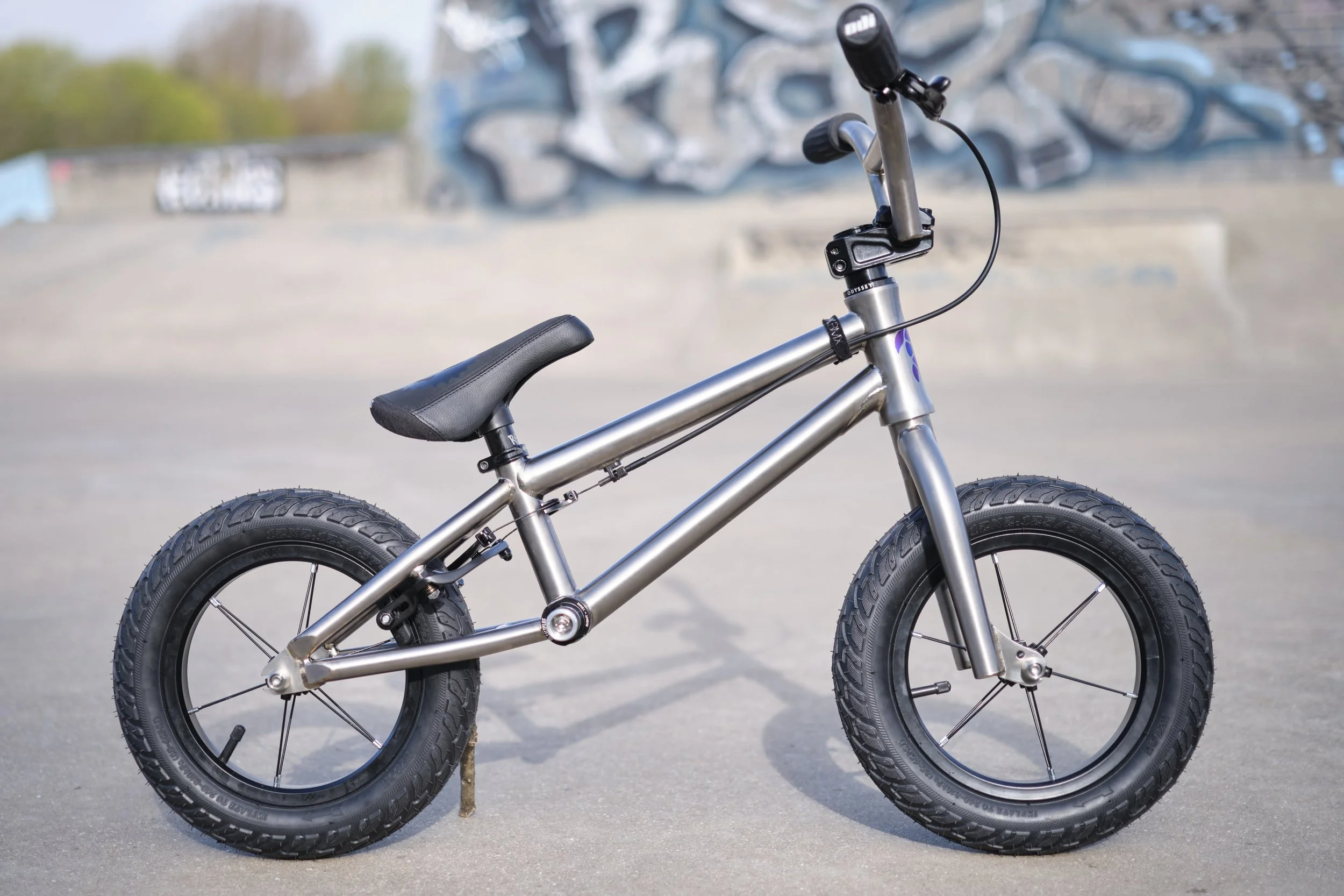 Silver children's balance bike with black tires and seat, outdoors on pavement with a graffiti-covered wall in the background.