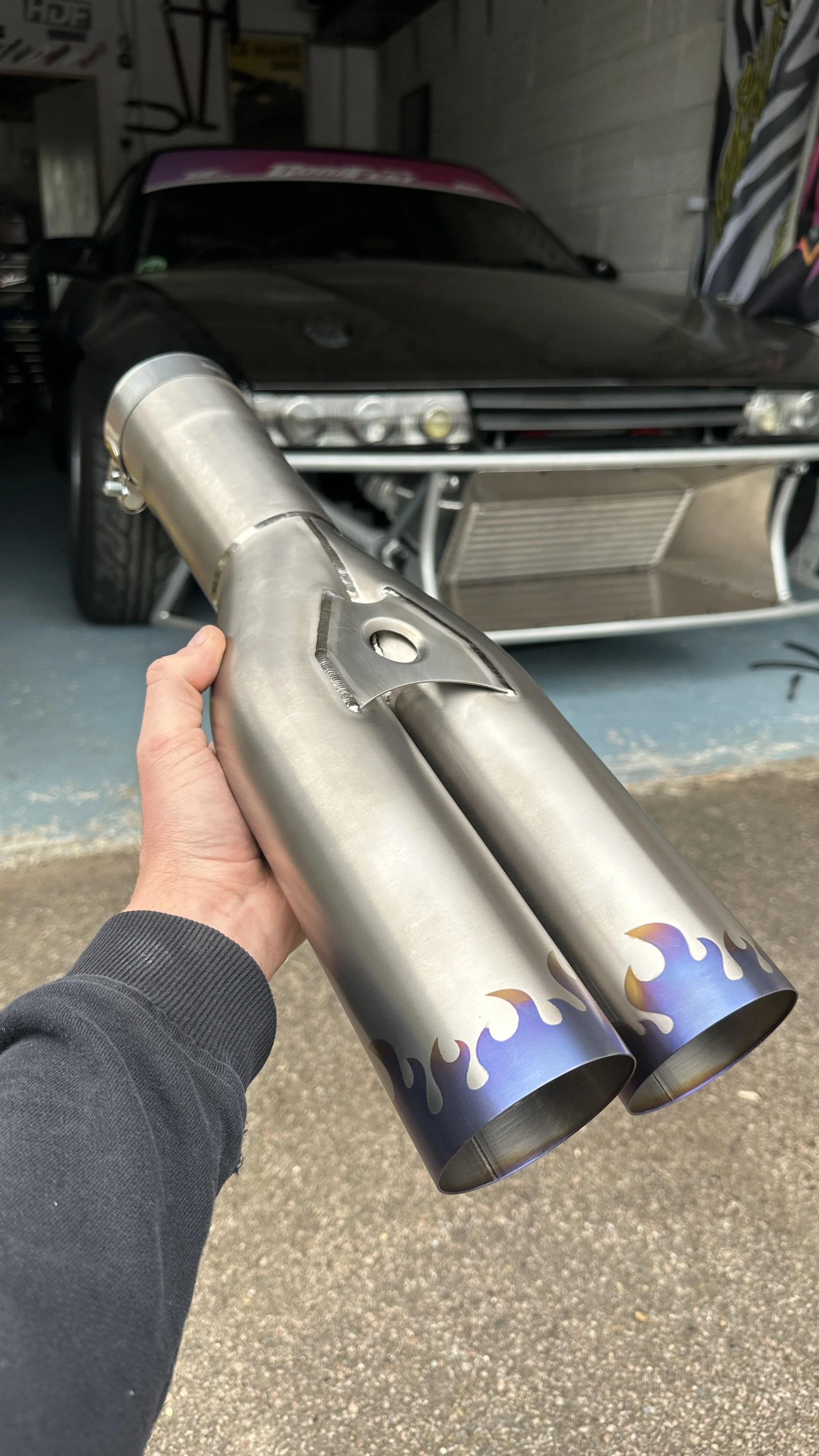 A person holding a two-exit exhaust pipe with flame decals in front of a black car with a prominent front grille and bumper inside a garage.