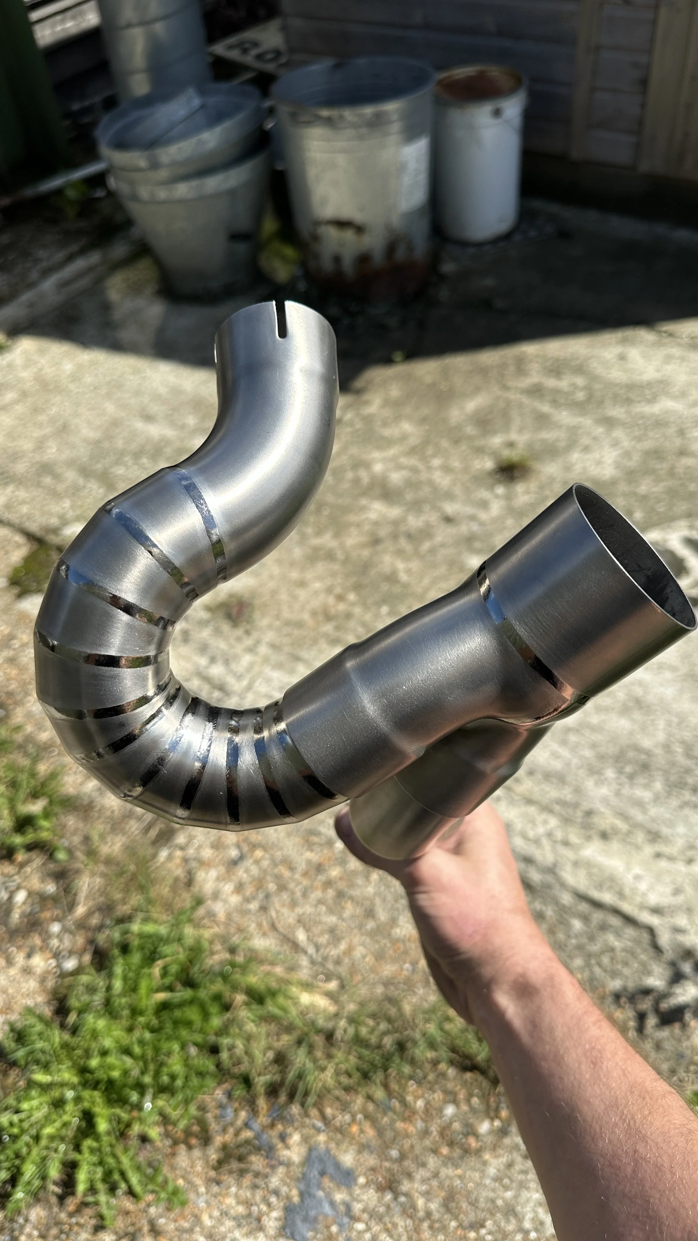 A person holding a shiny metallic pipe with a curved section and a straight section, outside on a concrete surface with several rusted and weathered metal barrels in the background.