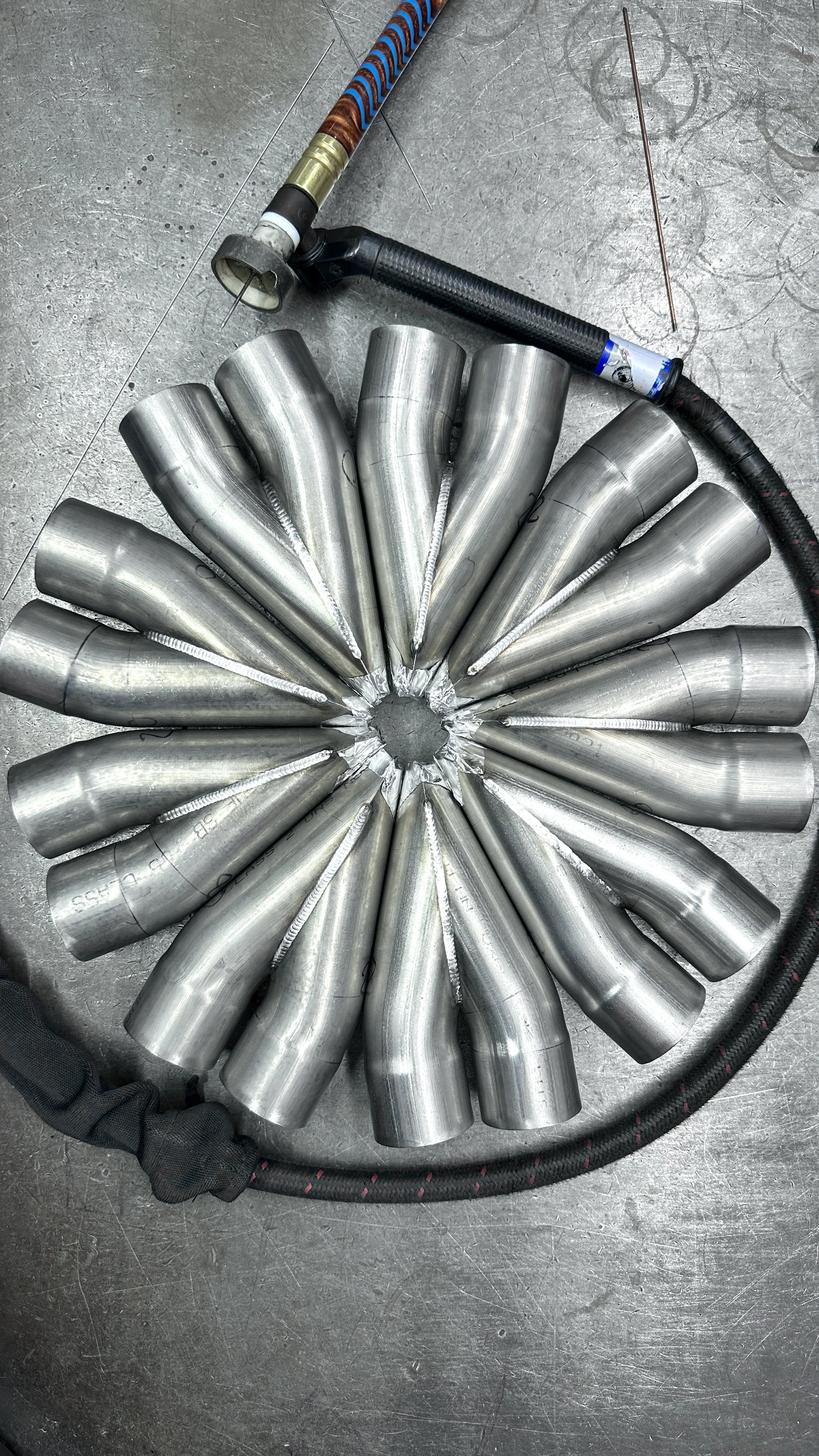 A circular arrangement of welded metal pipes resembling a flower on a metal work surface with welding tools and cables around.