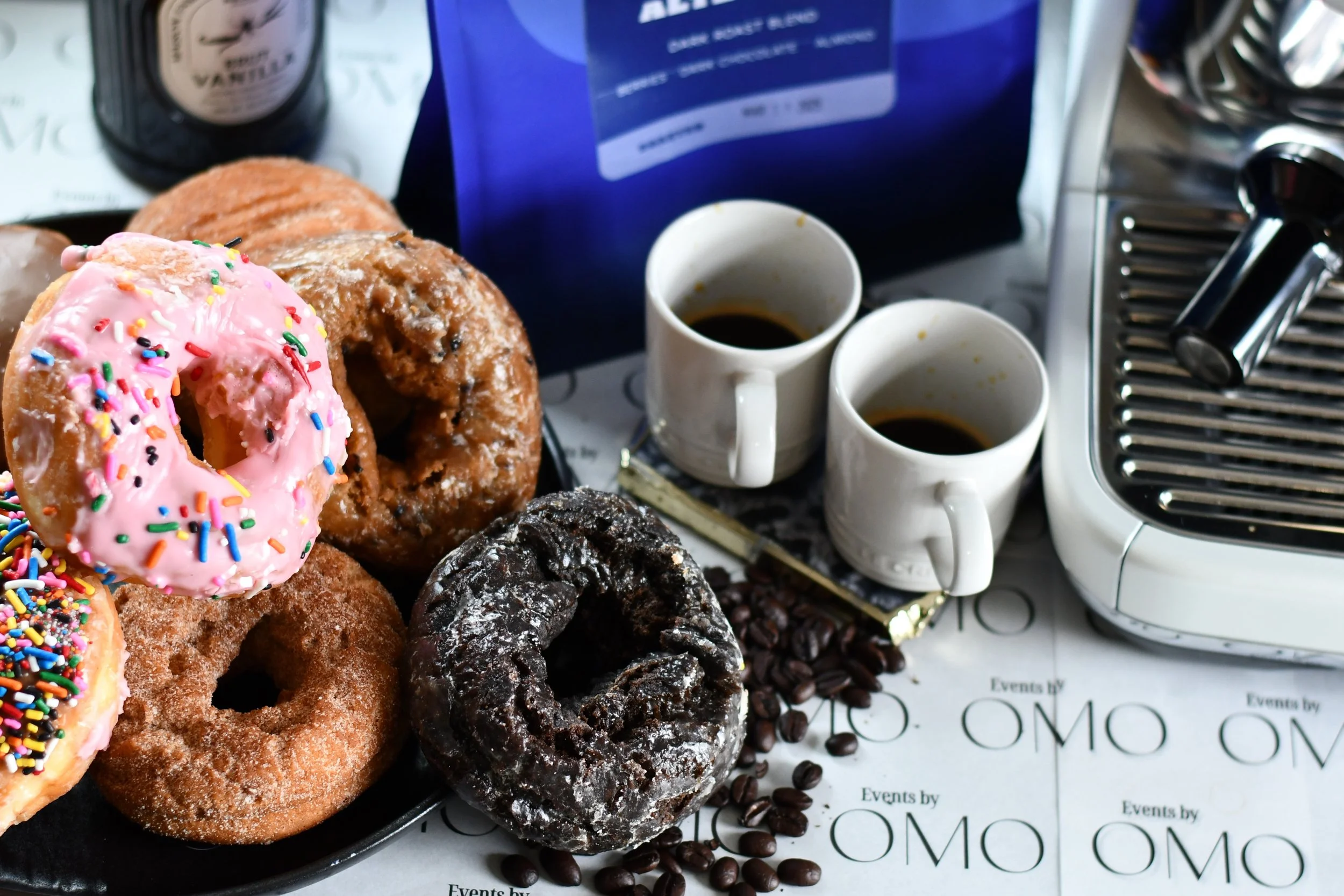 Assorted donuts with pink frosting and sprinkles, chocolate glazed, and plain, placed on a black tray. There are two cups of black coffee, coffee beans scattered on the table, and part of an espresso machine visible on the right.