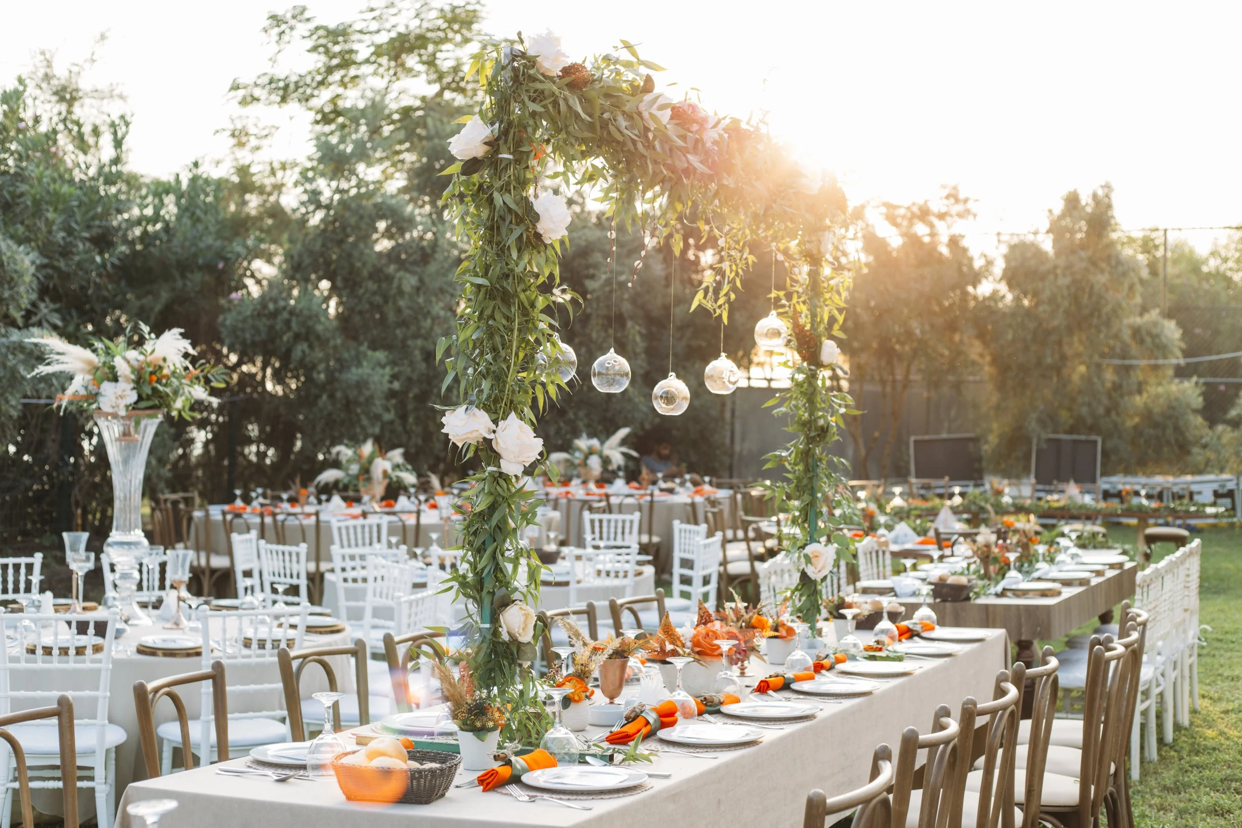 Outdoor event setup with decorated tables, chairs, and hanging glass orbs illuminated by warm sunlight.