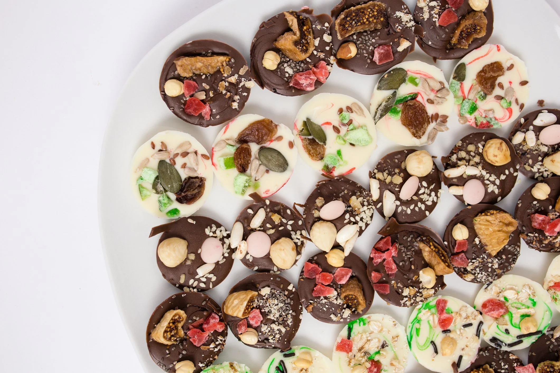 Assorted decorated chocolates on a white plate with various toppings like nuts, dried fruit, sprinkles, and colorful sugar decorations.