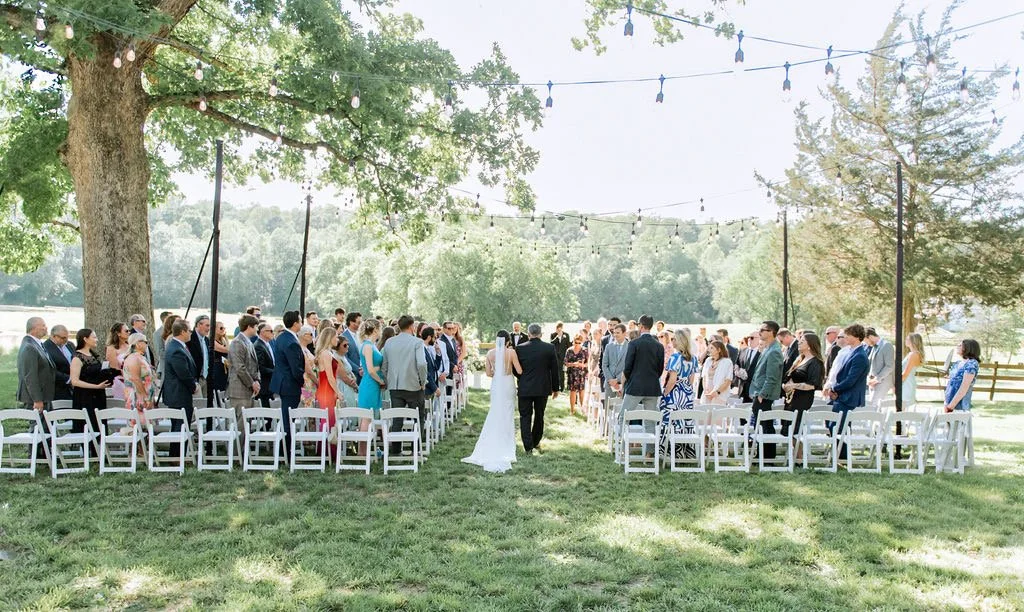 An outdoor wedding ceremony with guests standing on either side of the aisle, a bride walking down with her escort, under string lights on a sunny day in a lush green setting.