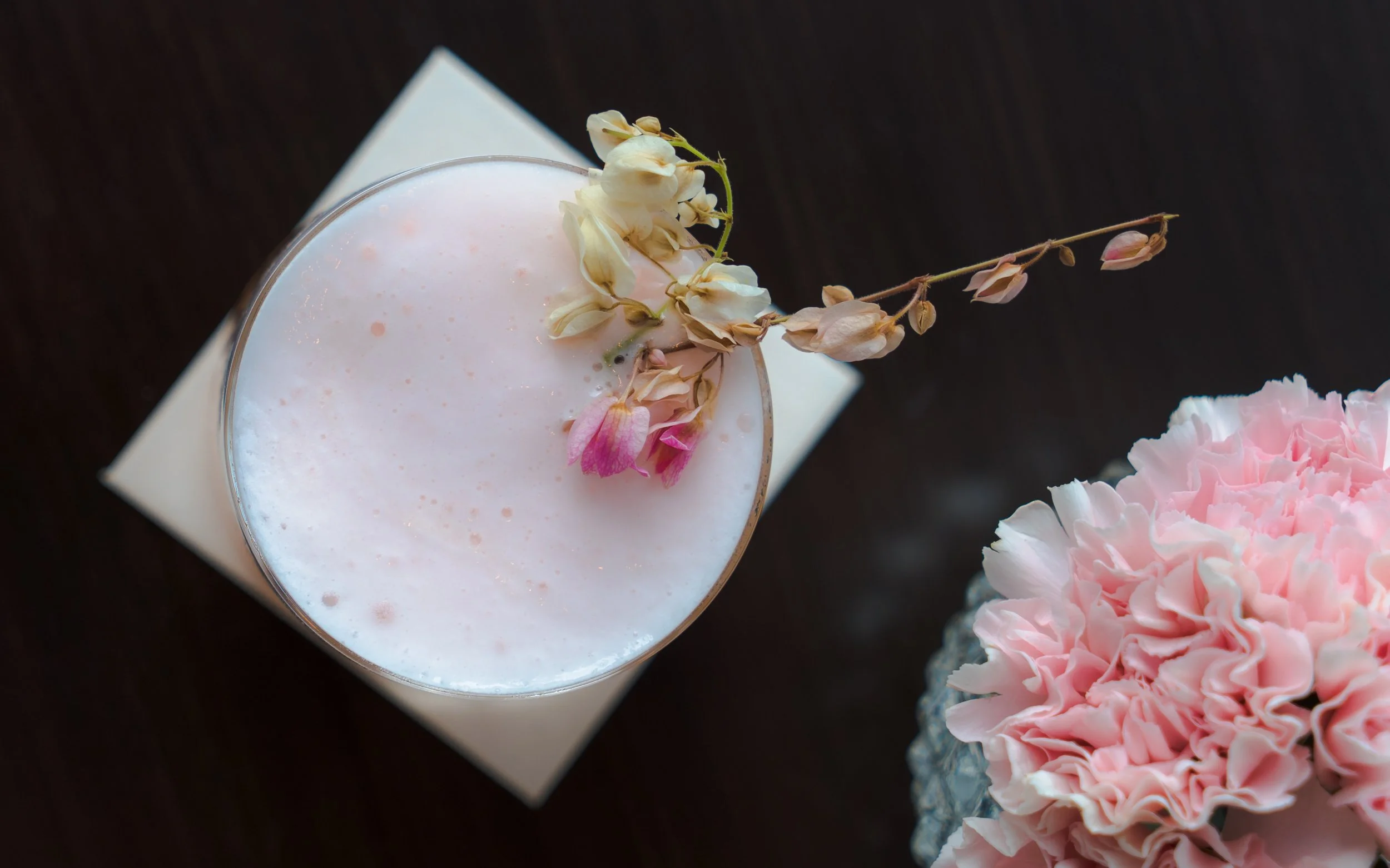 Pink frothy beverage in a glass with delicate dried flowers on top, placed next to a pink ruffled flower in a glass vase on a dark surface.
