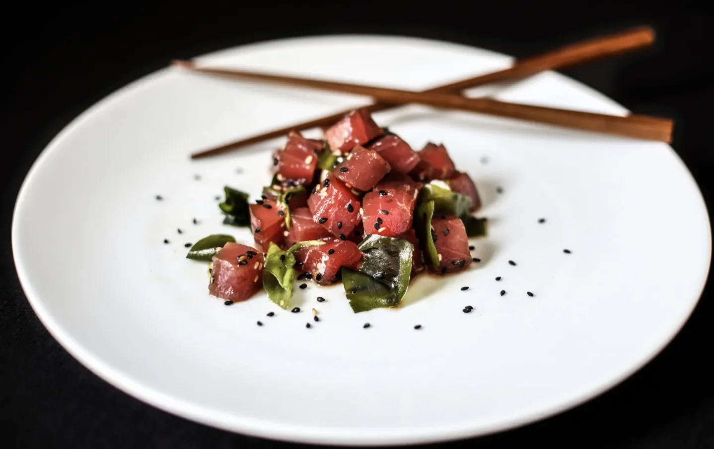 A white plate with chopped raw tuna garnished with black sesame seeds and green seaweed, with wooden chopsticks resting on the plate's edge.