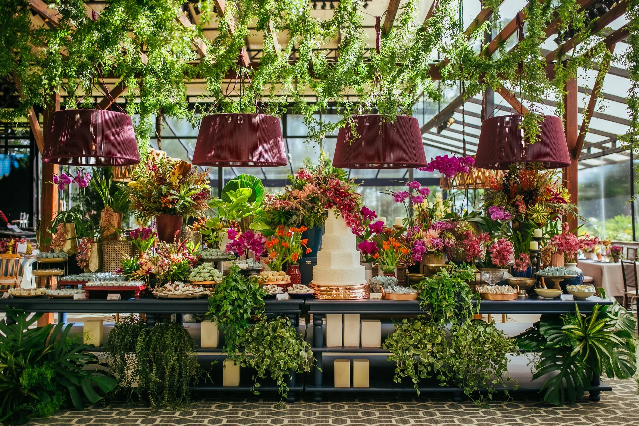 Decorated table at a celebration with a white tiered cake surrounded by various colorful flowers, green plants, pink lampshades, and food and dessert items on the table inside a glass conservatory.