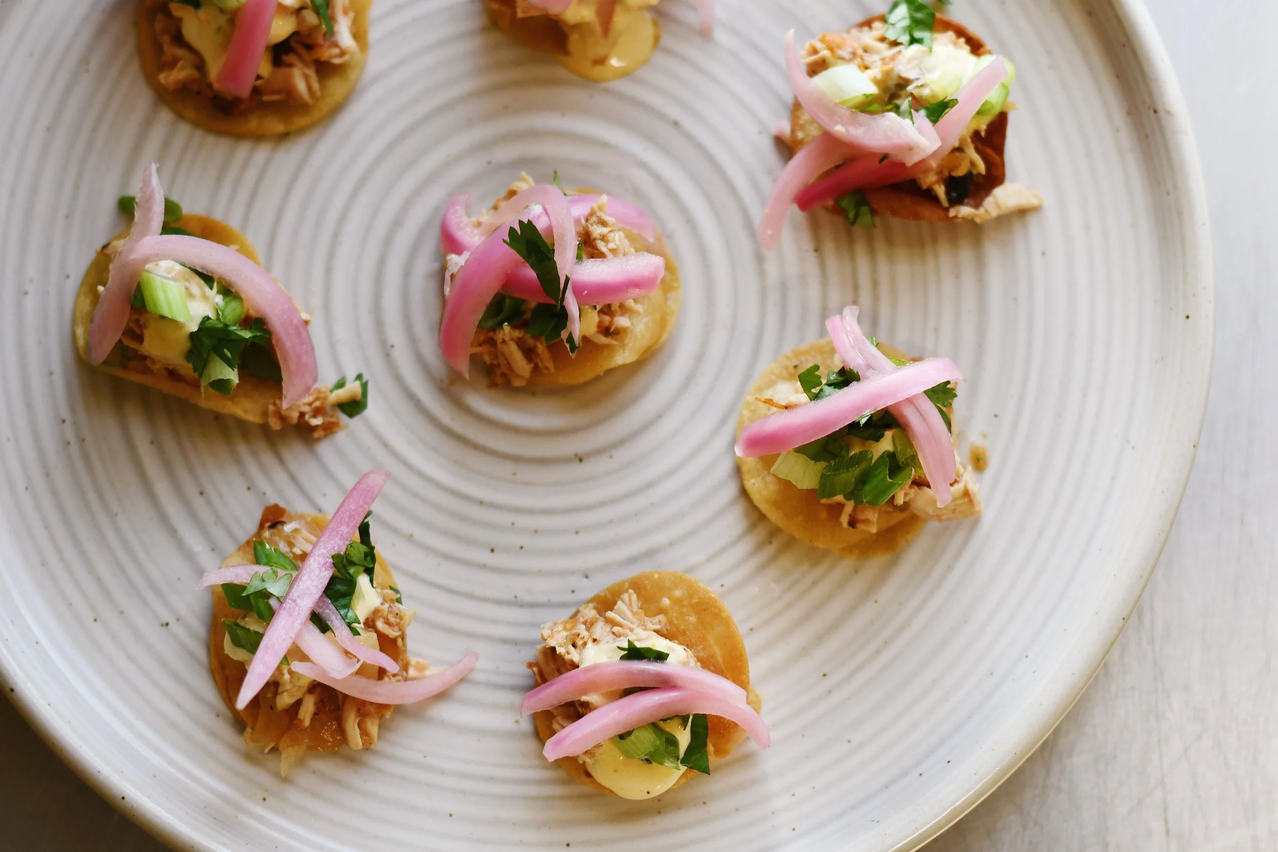Small appetizers with shredded chicken, chopped green onions, and pickled red onions on round corn cakes, arranged on a white plate.
