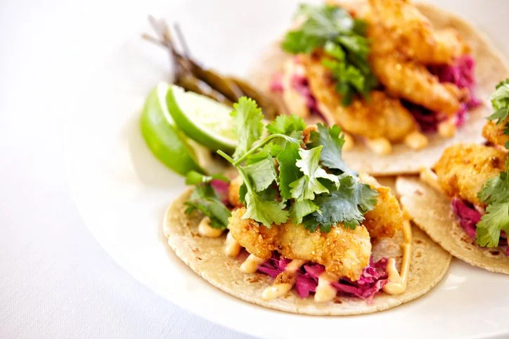 Three fish tacos with fried fish, purple cabbage, cilantro, and drizzled sauce on small corn tortillas, garnished with lime wedges and greens.