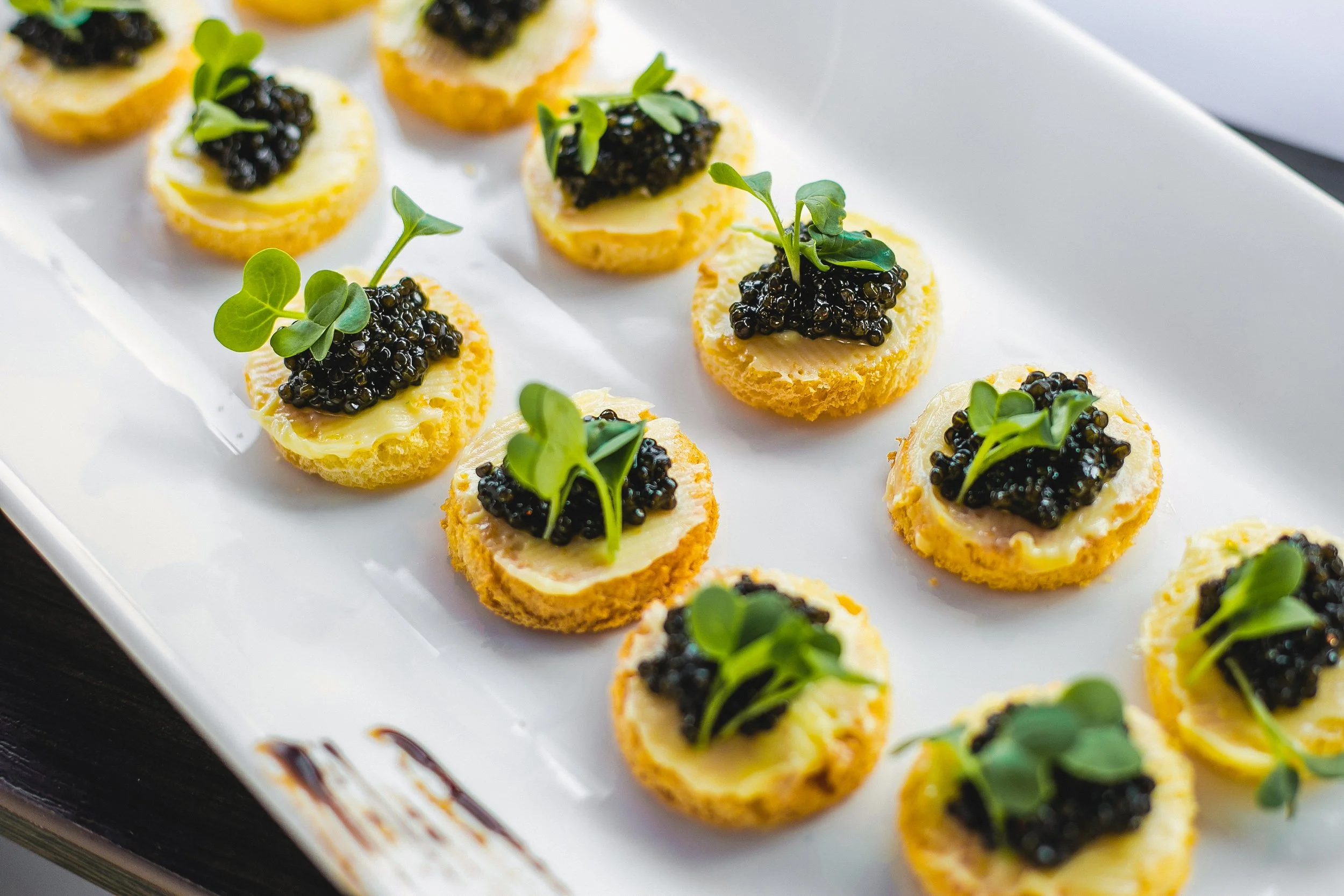 Canapé appetizers with a crispy base, topped with cheese, caviar, and microgreens on a white serving platter.