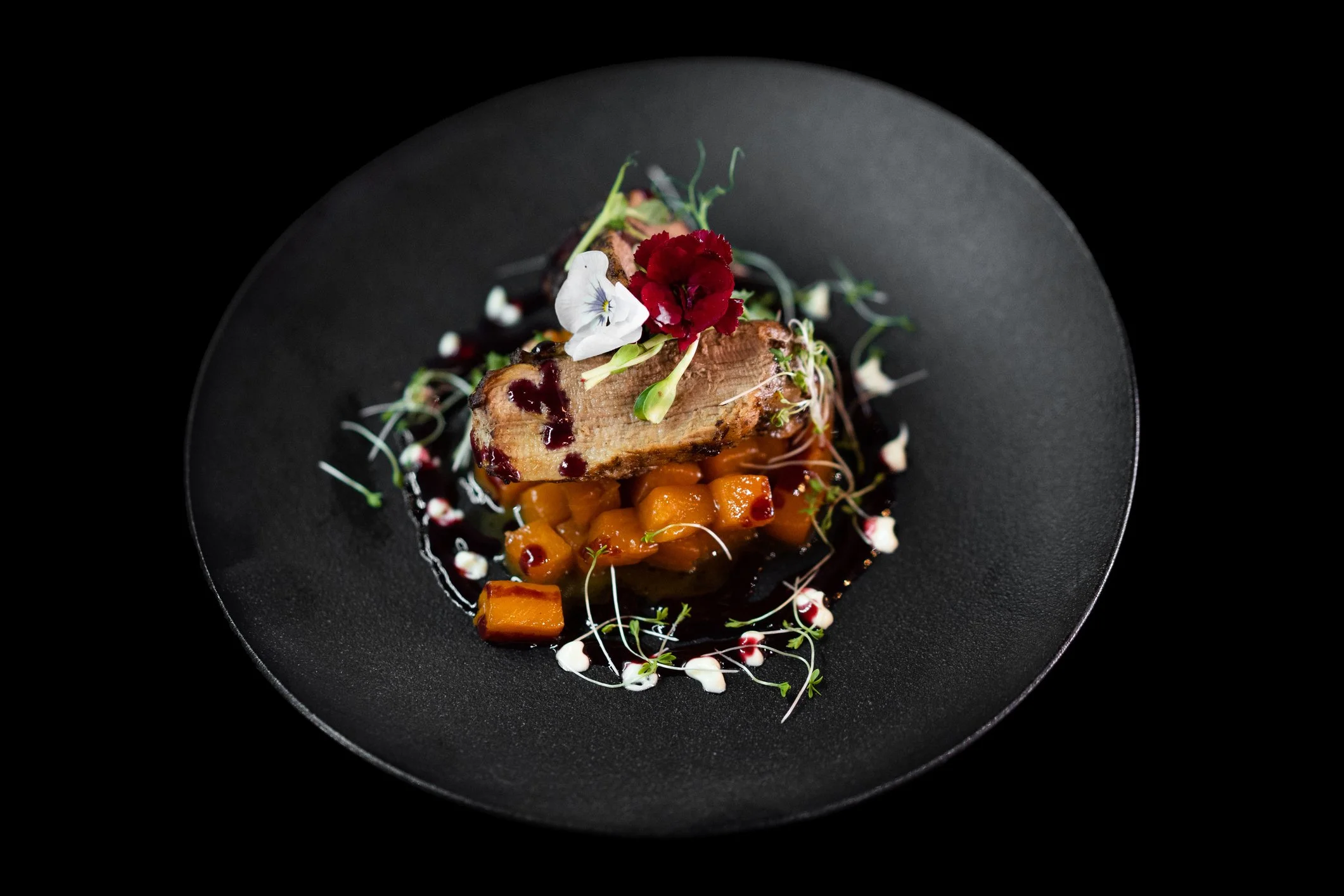 A gourmet plated dish on a black plate with a piece of meat, orange diced vegetables, edible flowers, microgreens, and a dark sauce.